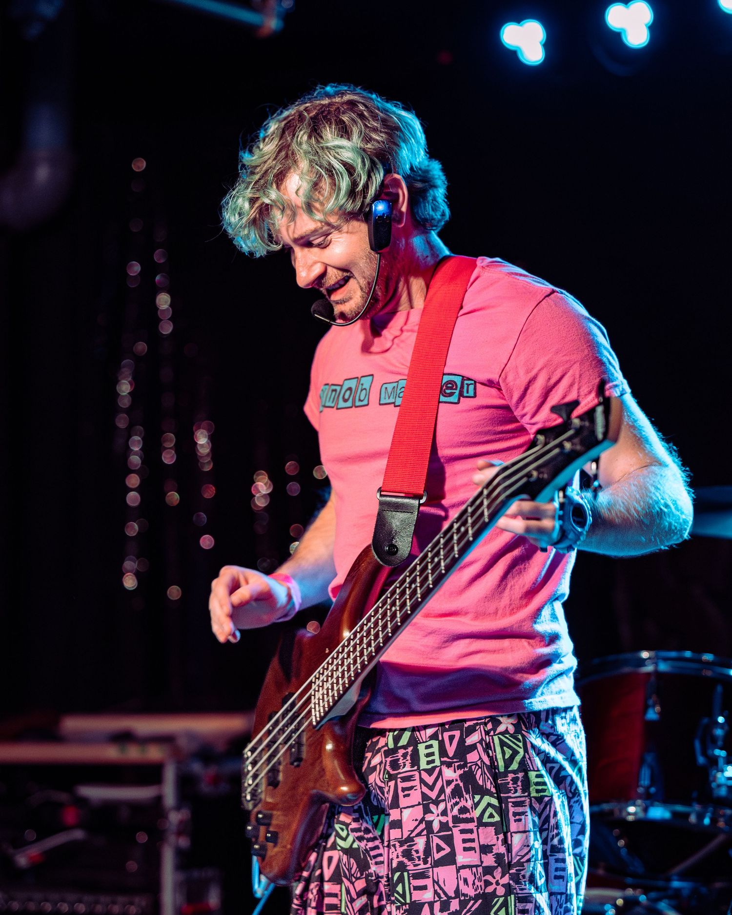 Male guitarist in pink shirt and patterned pants playing bass guitar on stage under lights.
