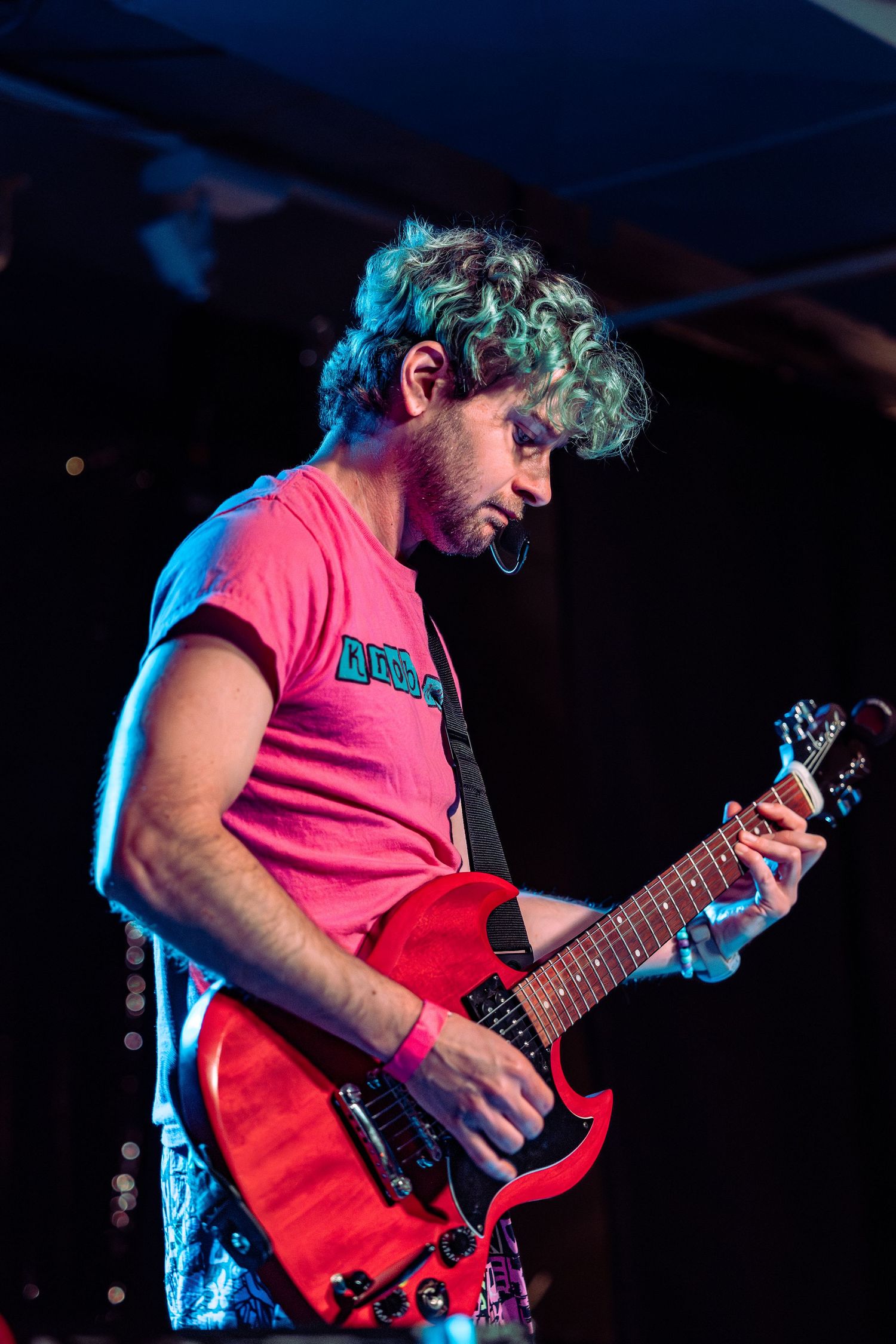 Curly-haired guitarist in pink t-shirt playing red electric guitar on stage under blue stage lights.