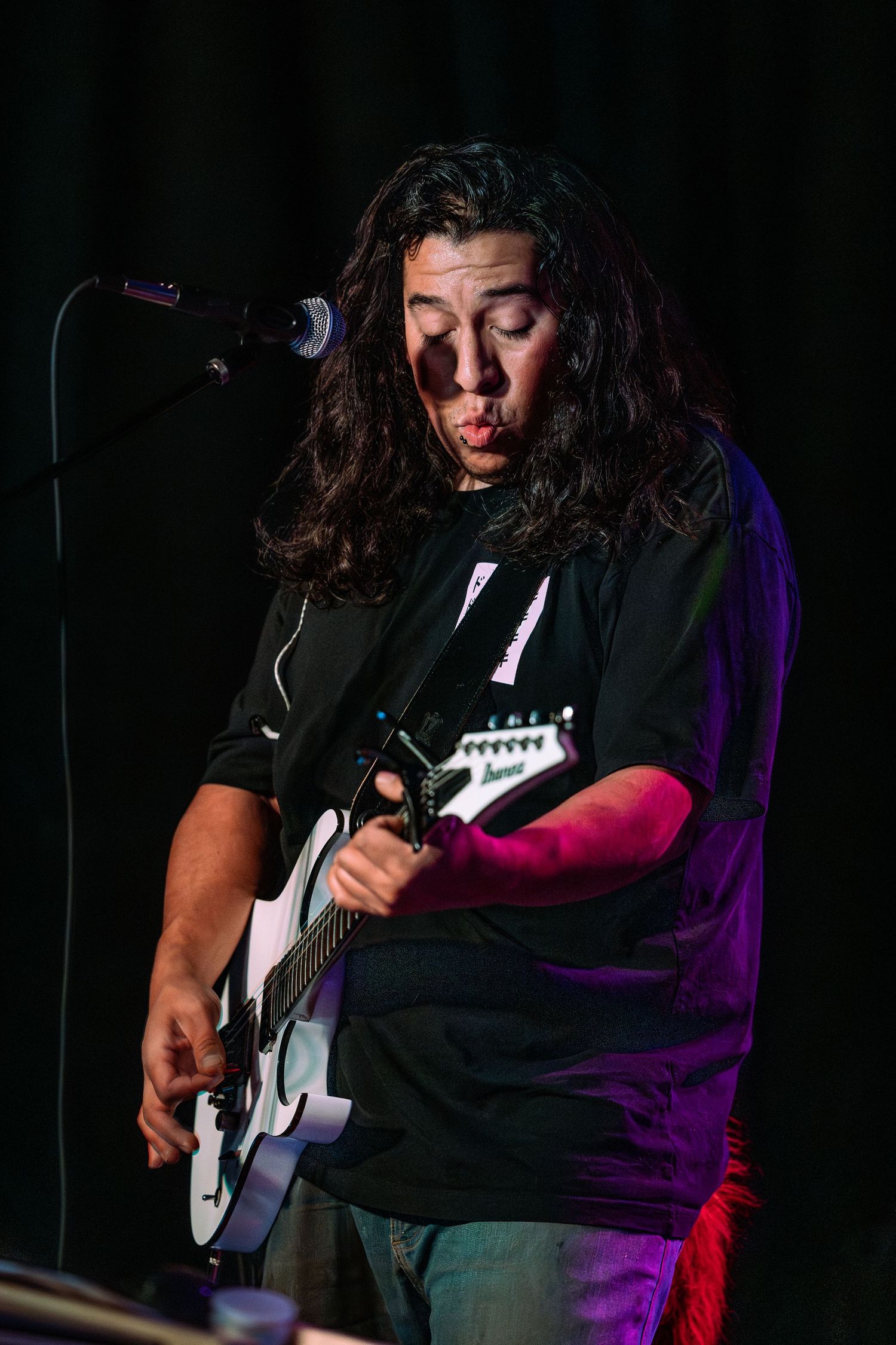 Male guitarist with long dark hair plays white electric guitar on stage under colorful stage lighting.