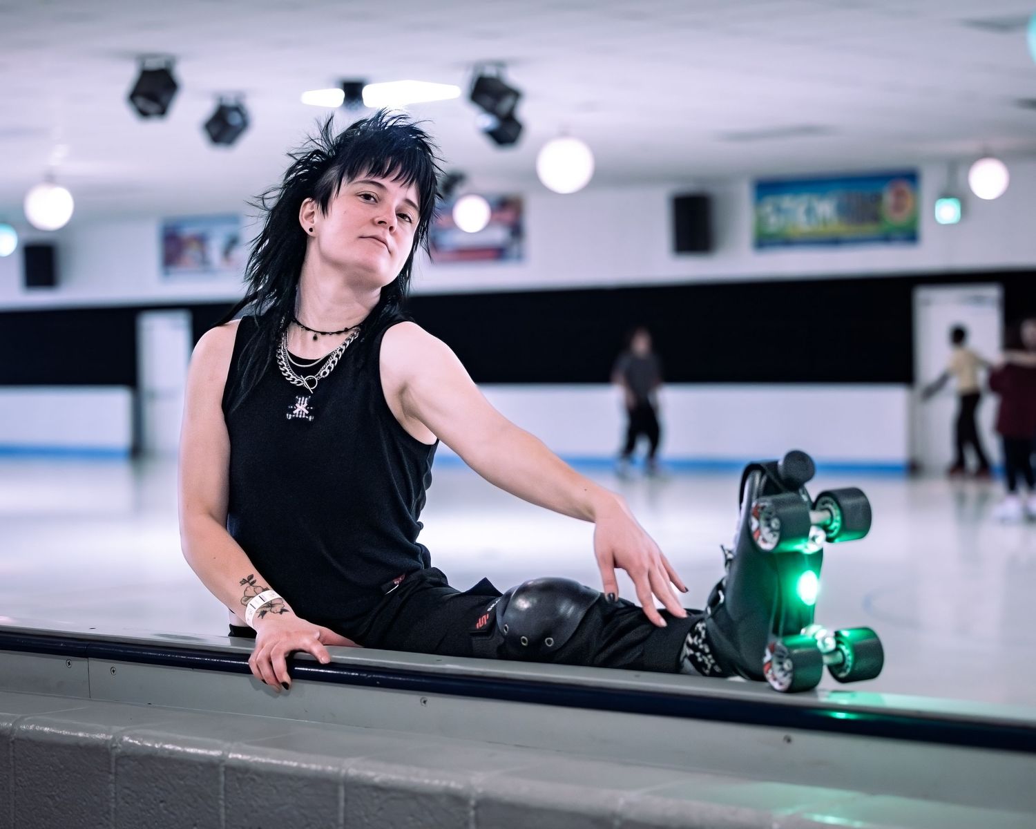 Person with black mullet hair in roller skates at an indoor skating rink, wearing black sleeveless top and accessories.