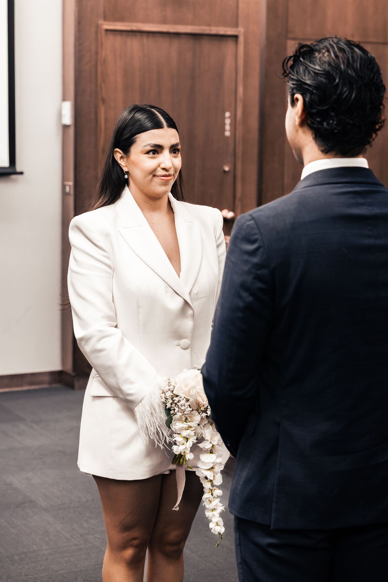 Bride in white blazer dress holds floral bouquet, exchanging vows with groom in dark suit at courthouse wedding.