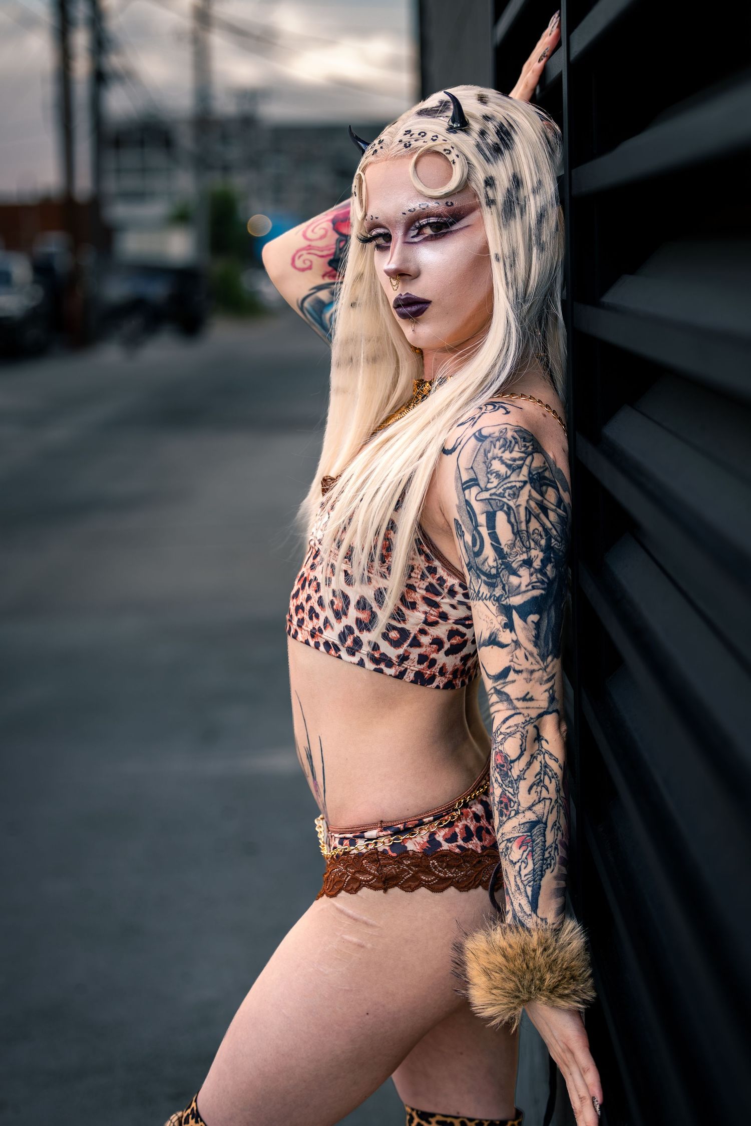 Tattooed blonde woman in leopard print crop top with horn makeup leaning on wall.