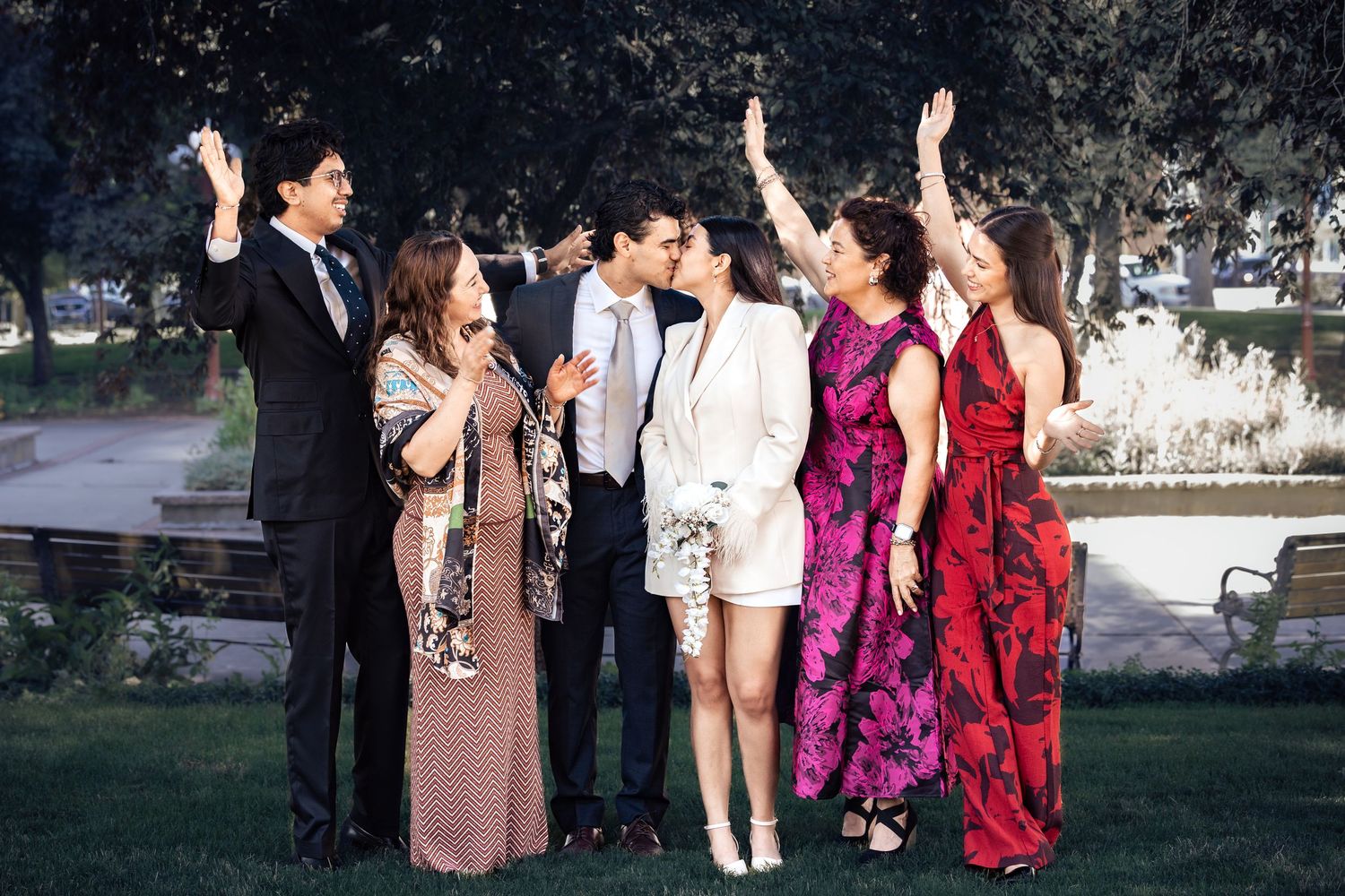 Wedding party celebrating outdoors in a park, guests in formal attire cheering as couple kisses.
