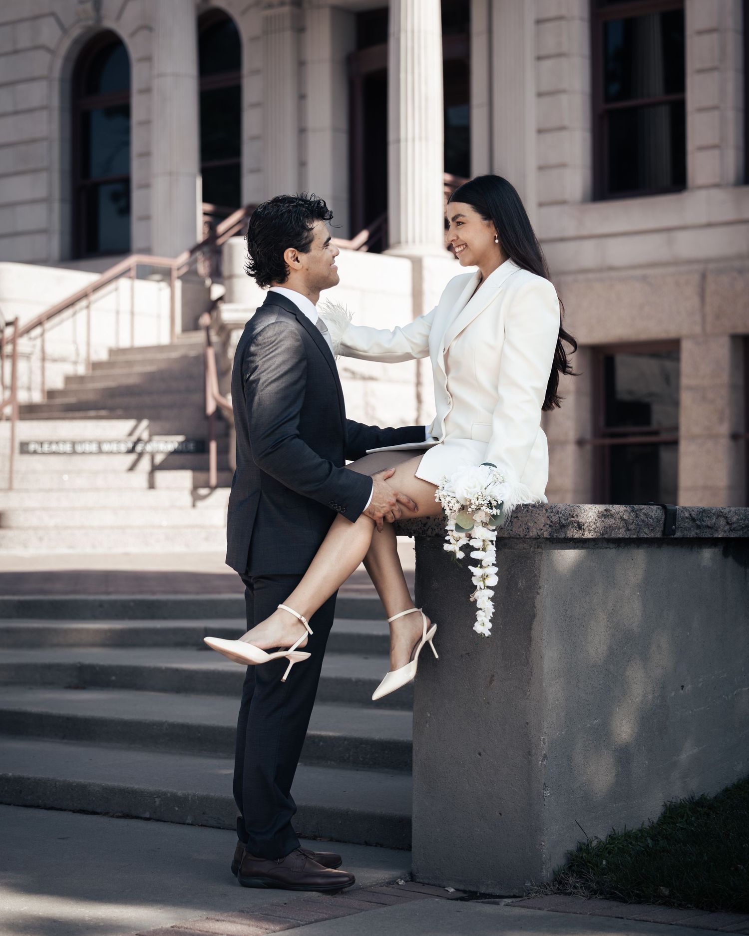 Couple in formal attire sharing a romantic moment outside a courthouse, woman in white blazer dress seated on stone ledge.