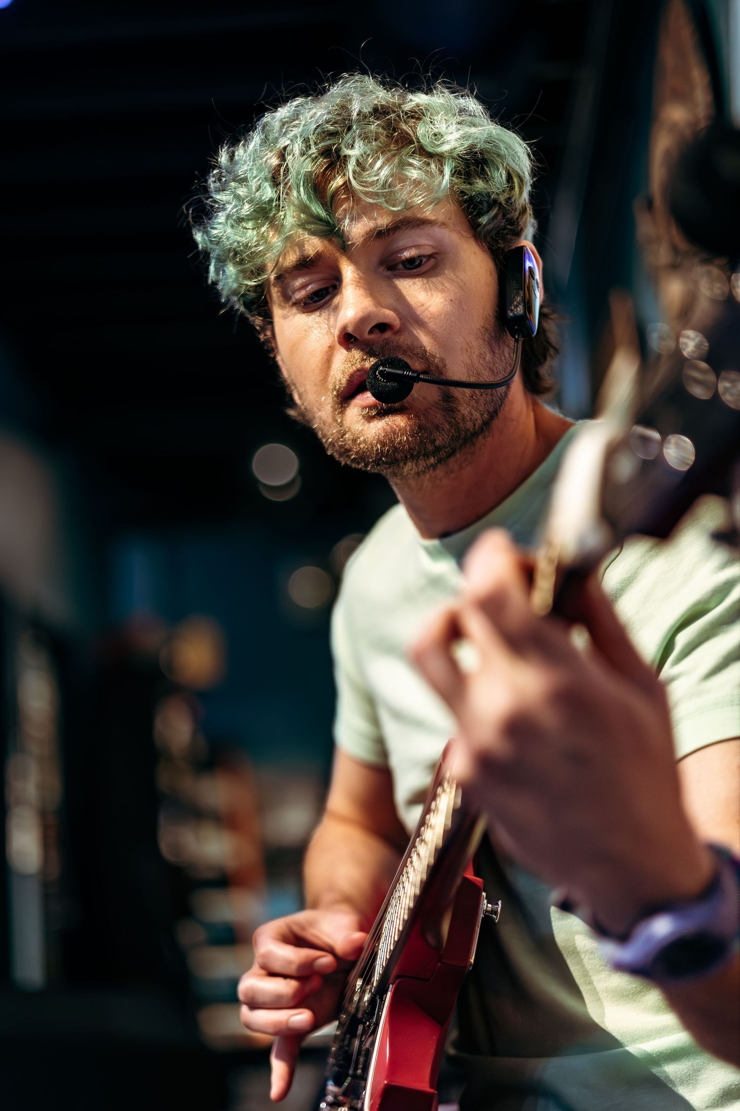 Musician with curly green hair performs passionately while playing guitar and singing into a microphone on stage.