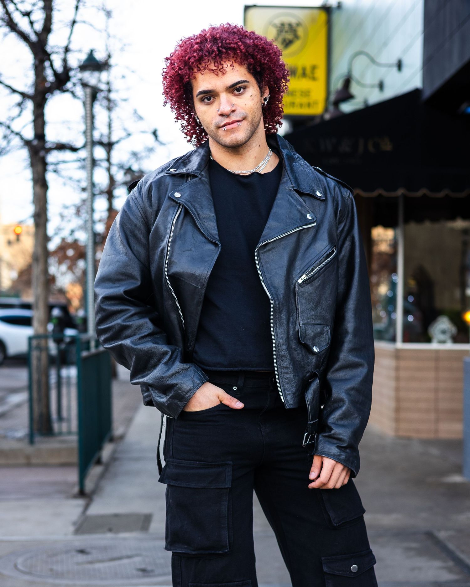 Urban fashion portrait with vibrant magenta curls and black leather jacket against storefronts.