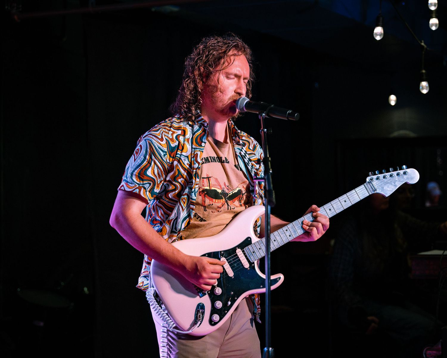 Blond guitarist in a colorful shirt playing a white Stratocaster guitar on stage.