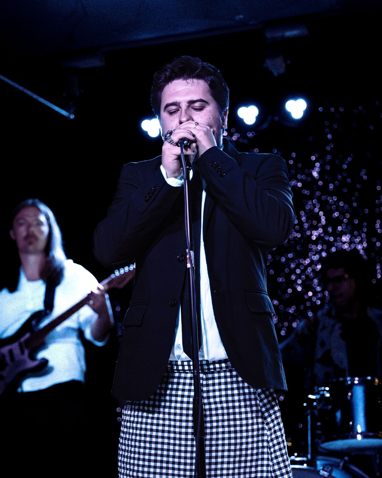 Male vocalist in black blazer and checkered pants performs at microphone on dark stage.