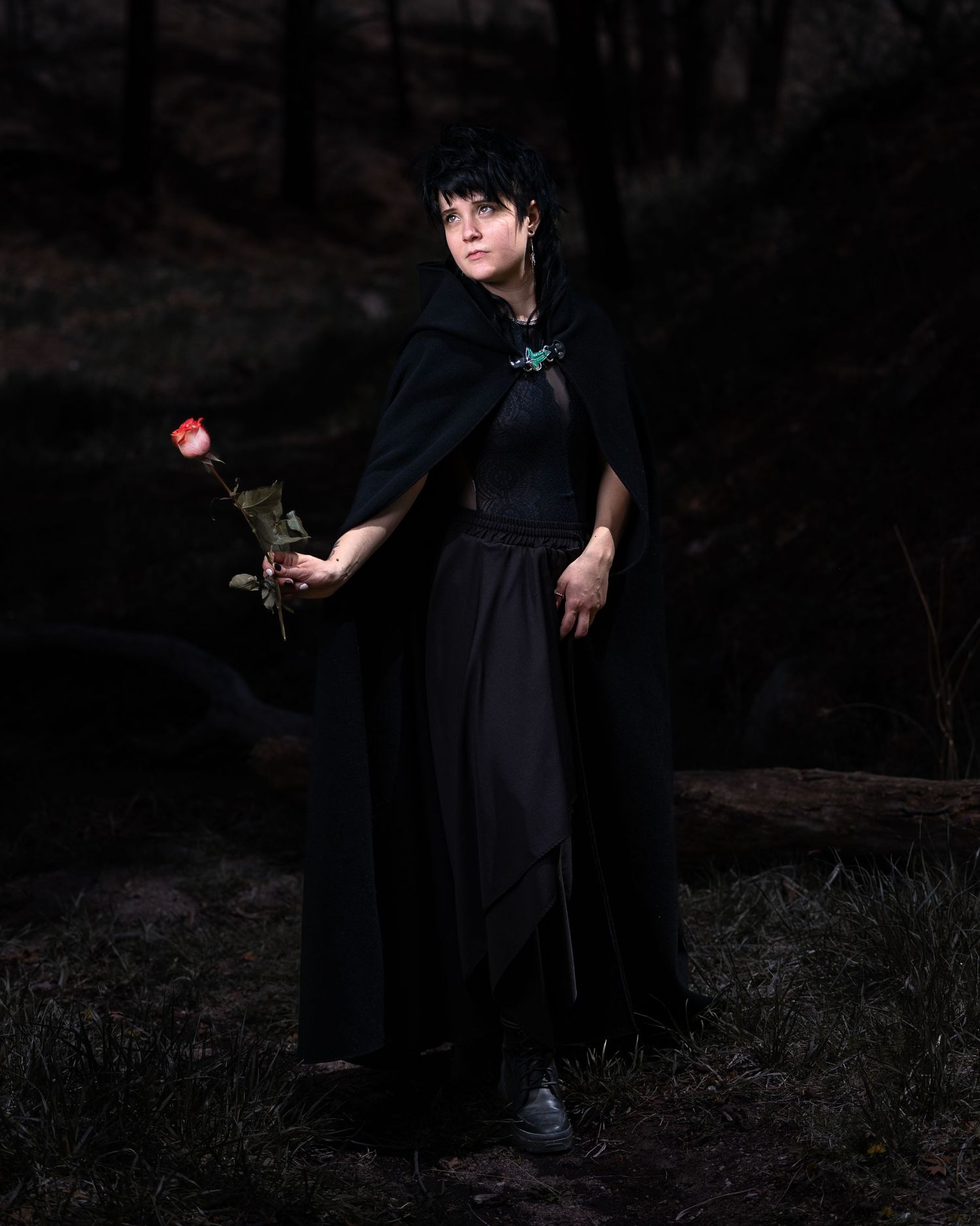 Woman in black cloak and dress holds red rose in dark forest, wearing turquoise brooch and white sneakers.