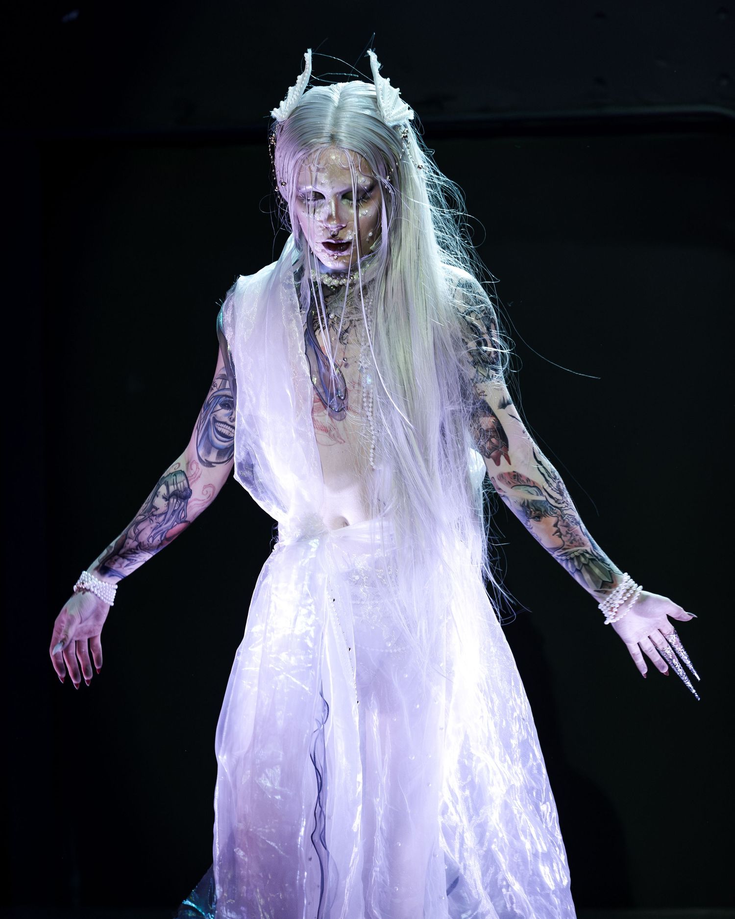 Tattooed performer in white sheer gown with horned headdress and long blonde hair on dark stage.