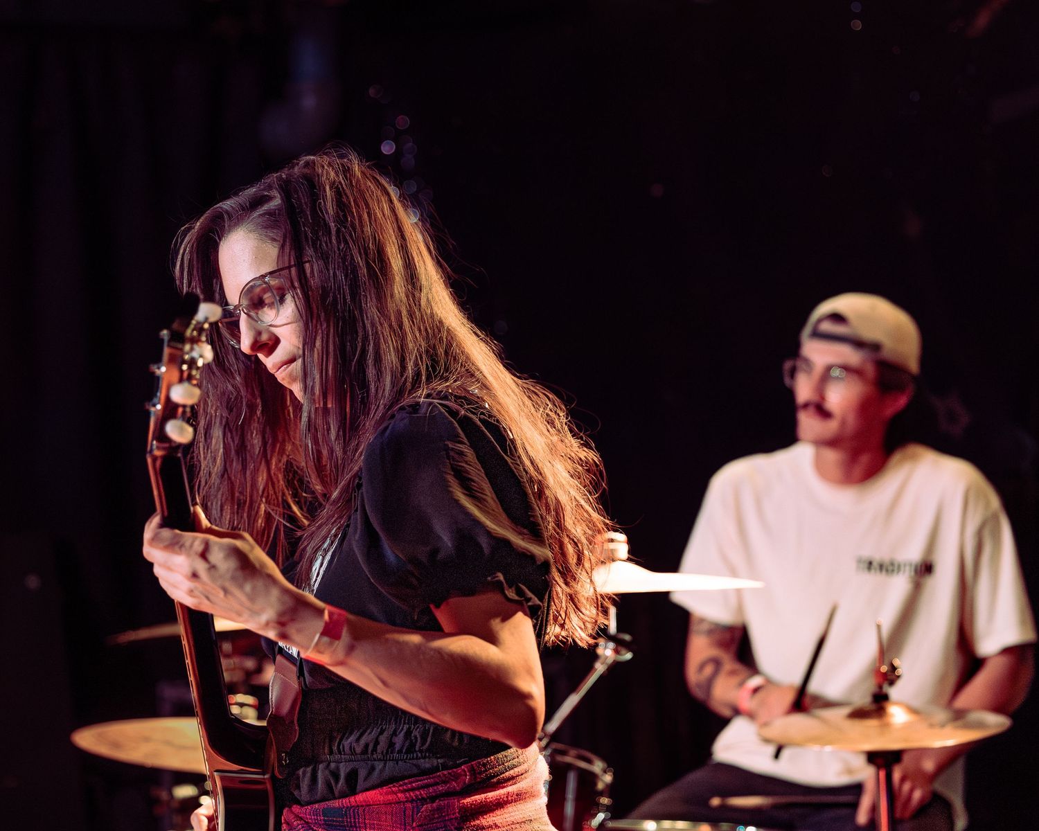 Woman playing guitar leaning forward with drummer visible in background on dark stage.