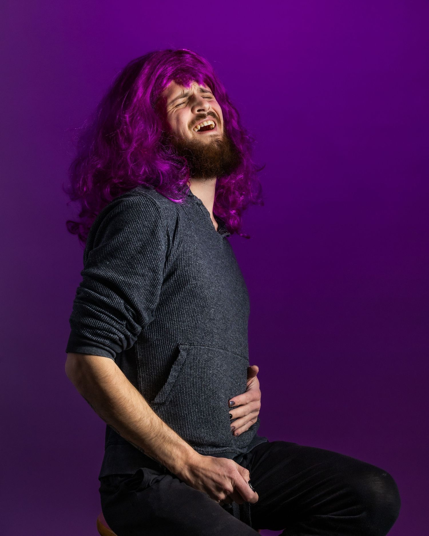 Person with long purple hair laughs joyfully while wearing a dark sweater against a vibrant purple background.