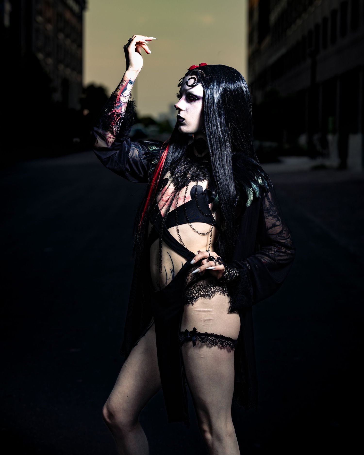 Tattooed gothic model in black lingerie and sheer robe posing on dark urban street at dusk.