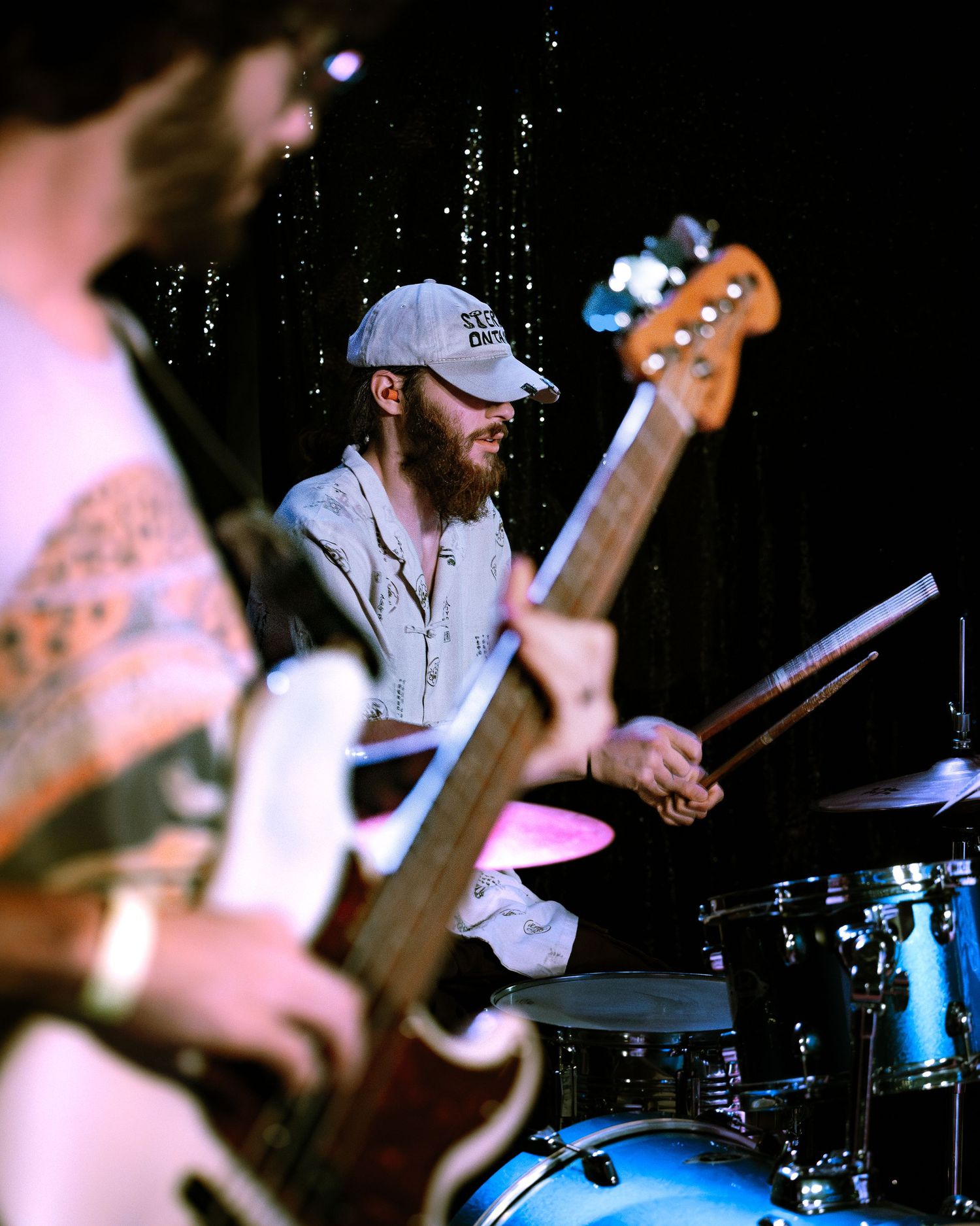 Bearded male drummer in cap plays drums on stage while guitarist performs in the foreground.