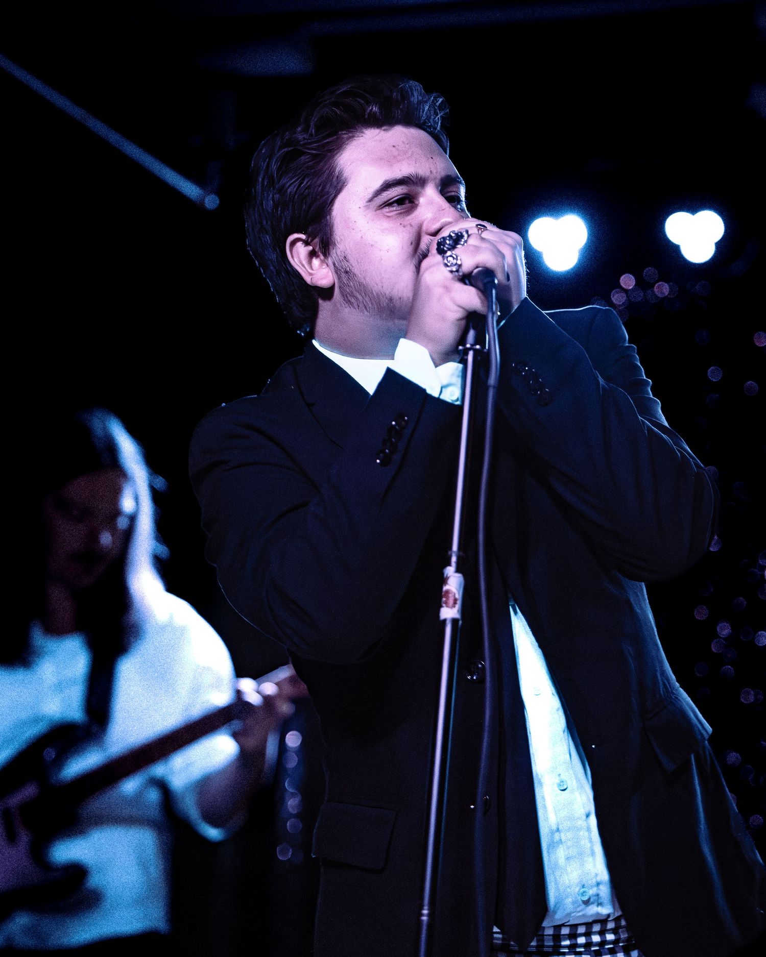 Male vocalist in black suit sings passionately into microphone under blue stage lights at concert.