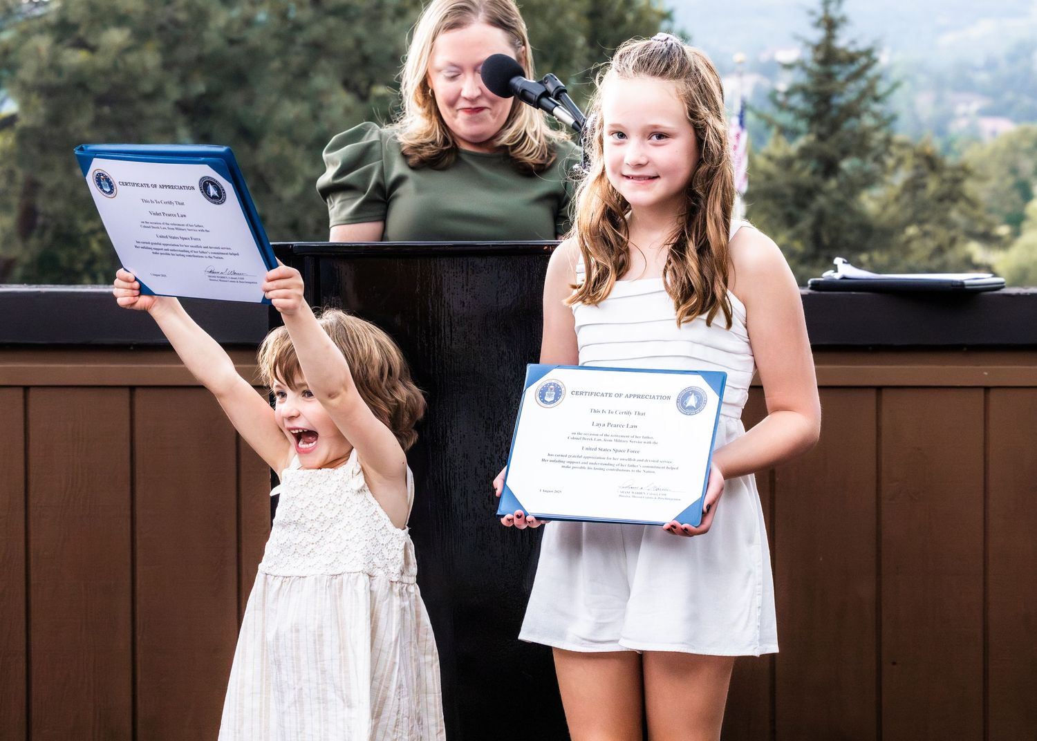 Children holding certificates during outdoor ceremony with mountain backdrop.