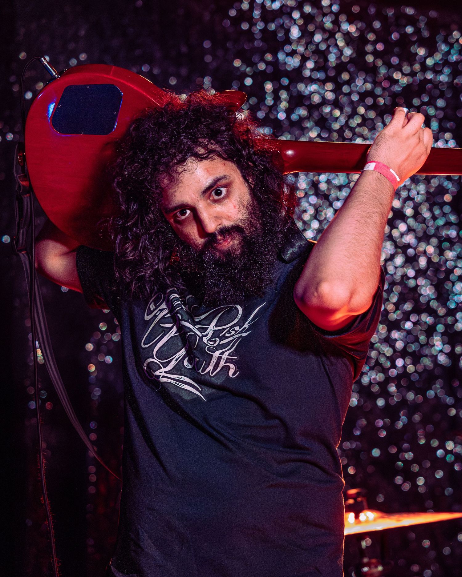 Bearded musician with curly hair balances red electric guitar on shoulder under stage lights.