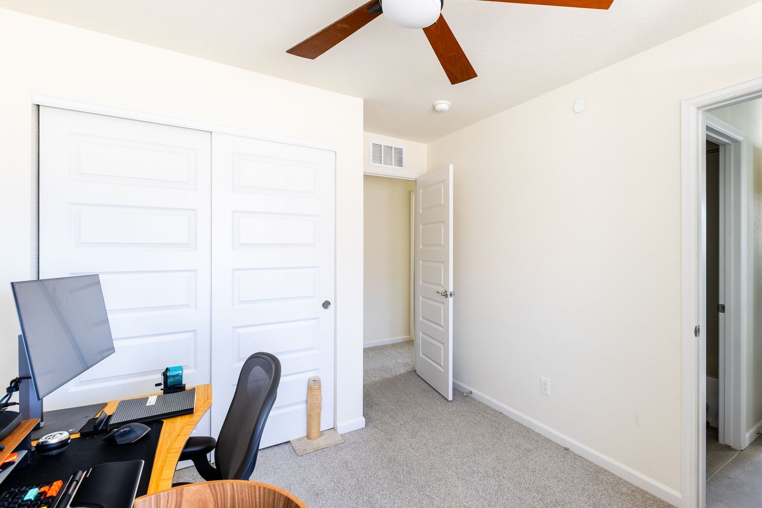 Bright home office with ceiling fan, white closet doors, desk with monitor, and carpeted floors in a neutral-toned room.