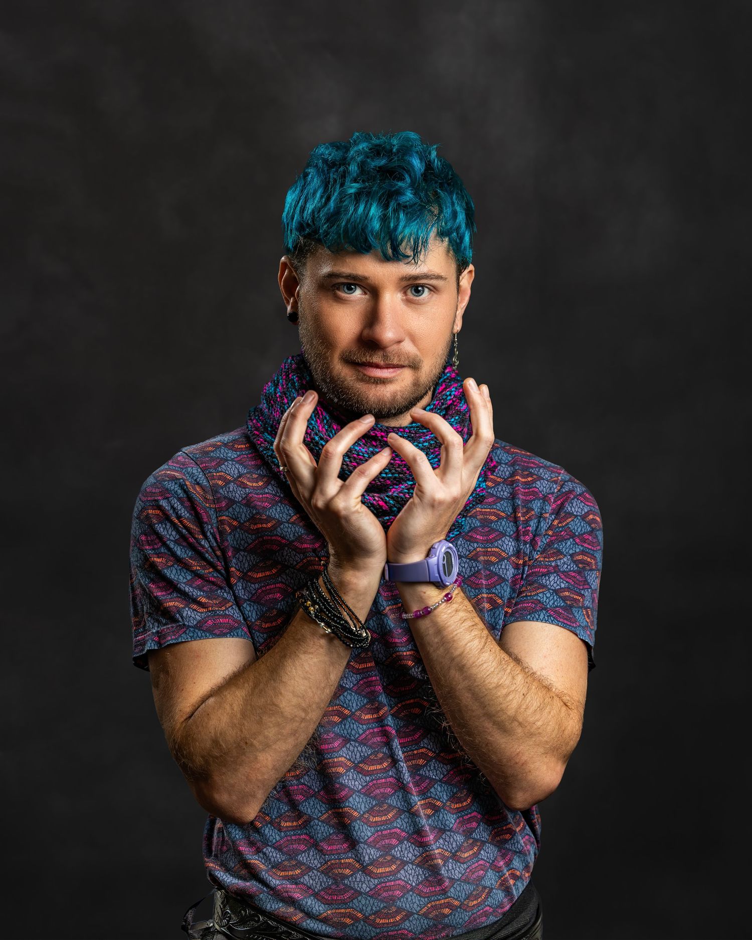 Man with vibrant teal hair poses with hands raised wearing a colorful patterned shirt in studio.