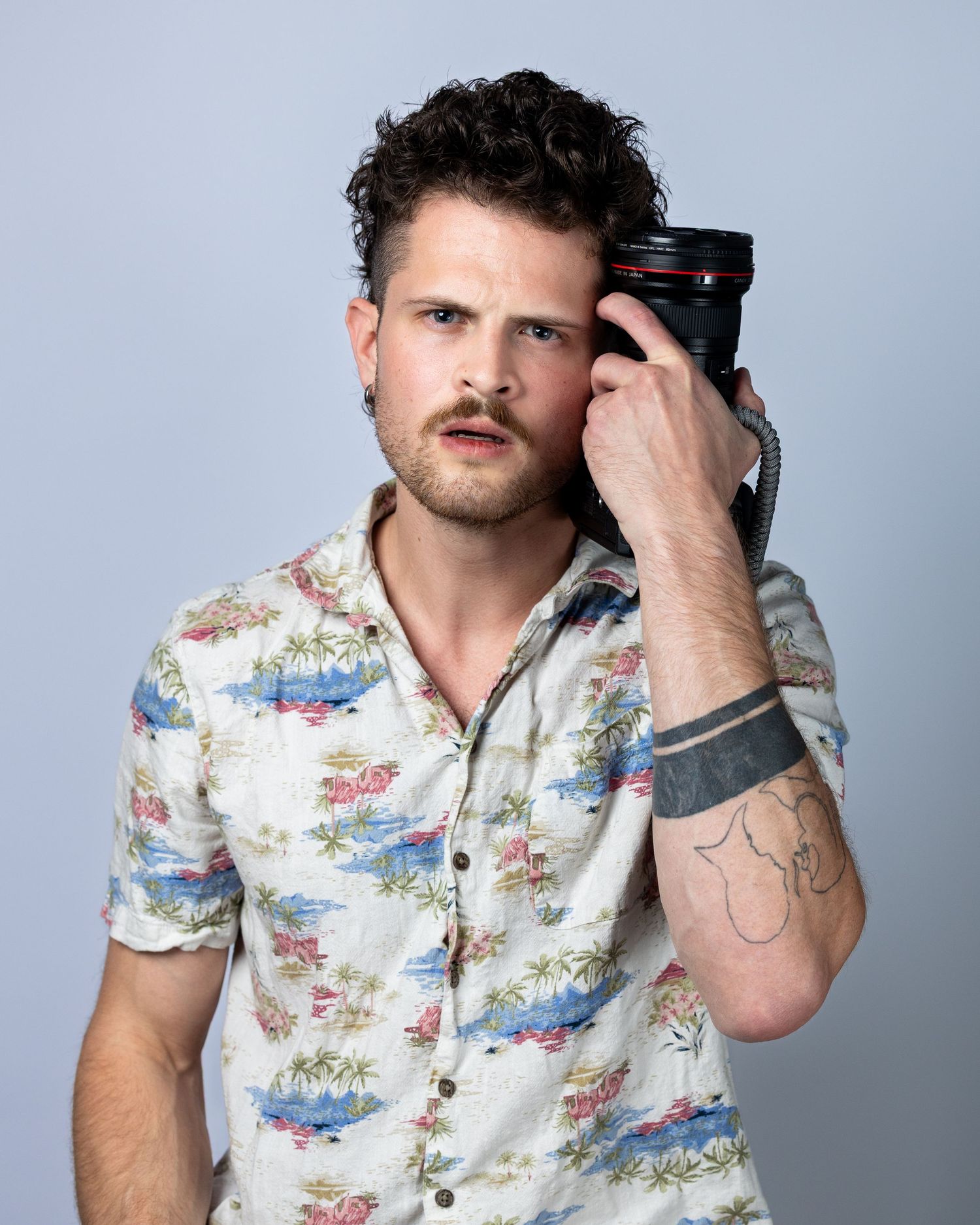 Young man with curly hair and mustache holding a camera lens, wearing a floral shirt with a visible arm tattoo.