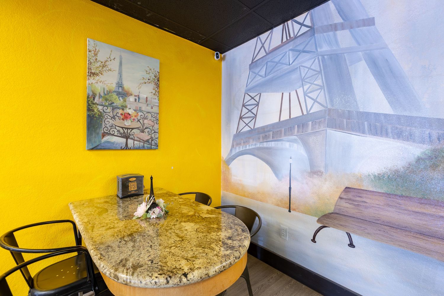 Cozy cafe corner with marble table, yellow walls, Eiffel Tower mural, and Parisian artwork creating a French-inspired dining ambiance.
