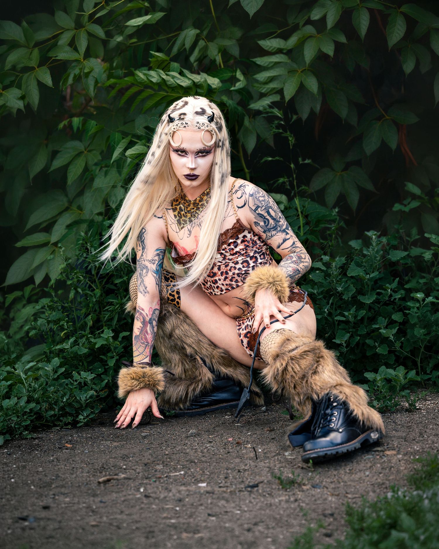 Tattooed blonde woman crouching in leopard print outfit with fur boots and dramatic makeup in forest.