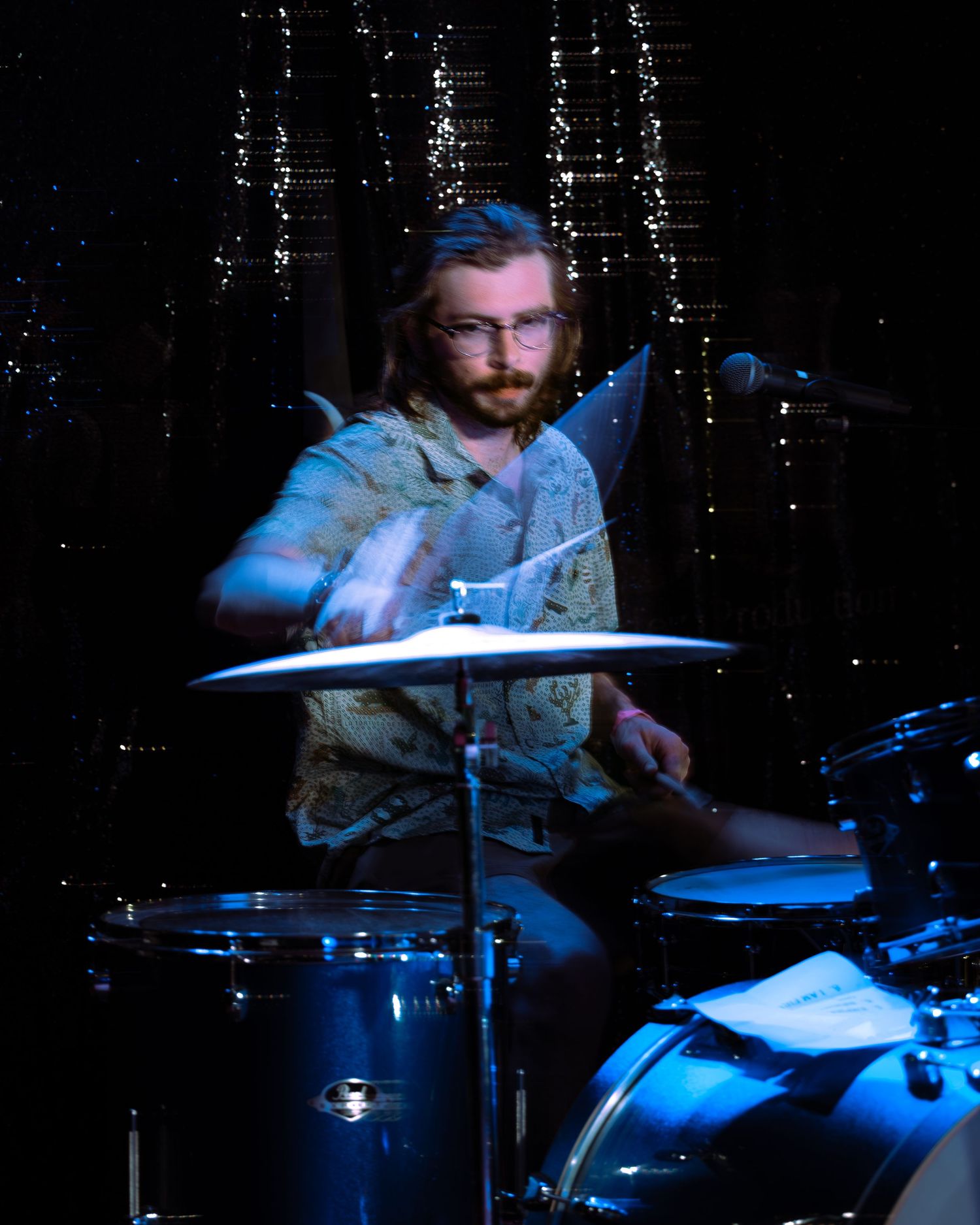 Bearded drummer with glasses playing Pearl drum kit under blue stage lights at concert.