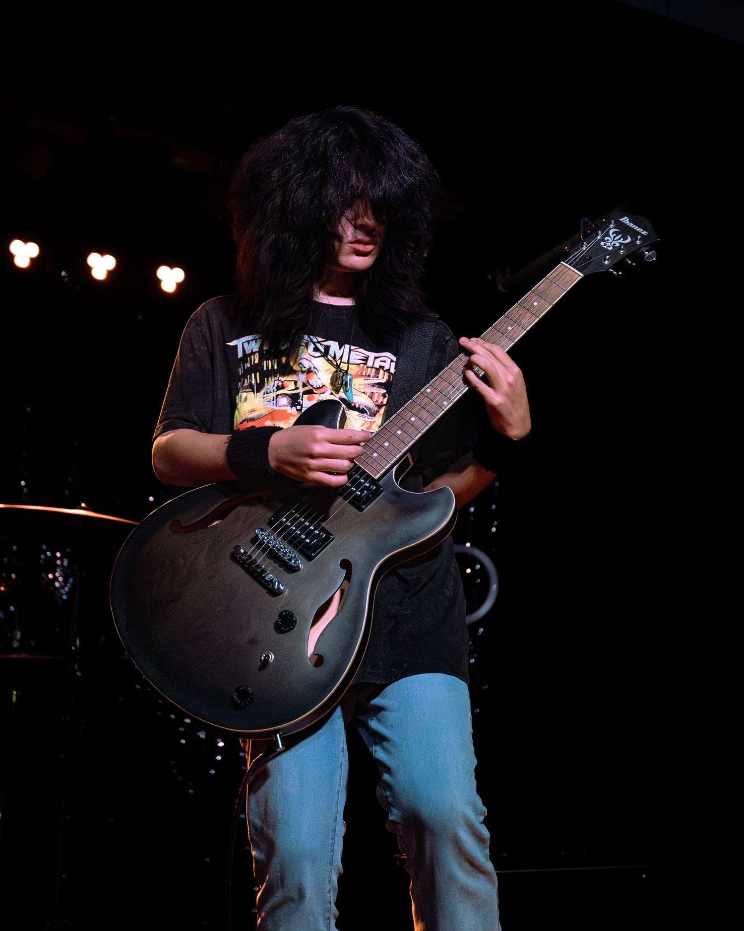 Young guitarist with long black hair playing dark electric guitar on stage in concert.