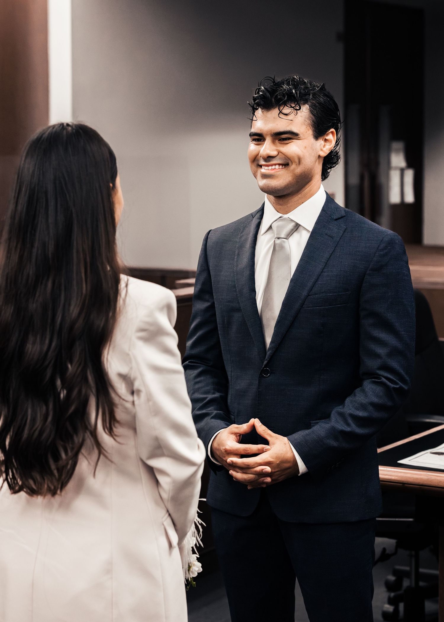 A smiling man in a navy suit with a gray tie converses with a woman in a white blazer in a modern office setting.