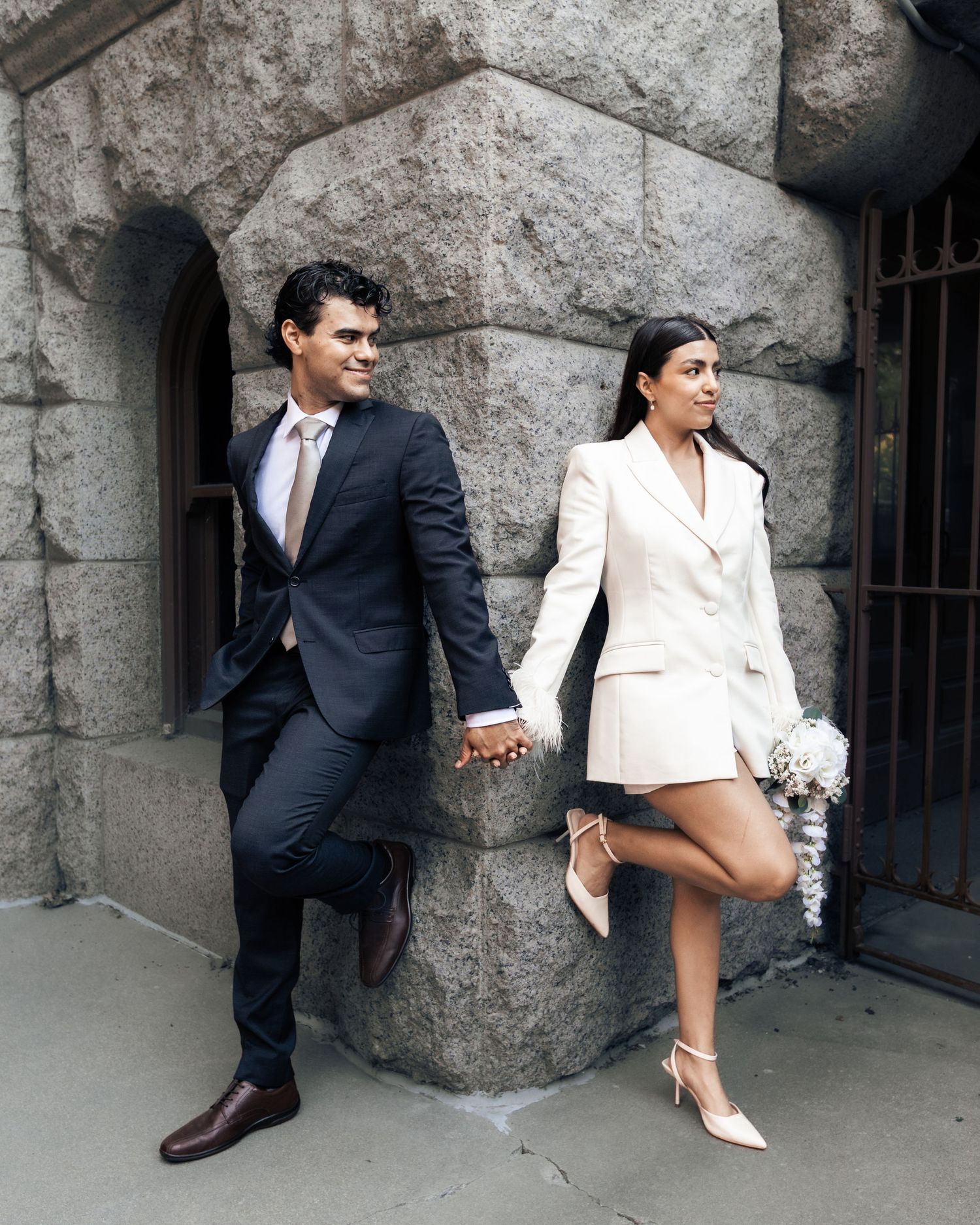 Couple in wedding attire holding hands outside stone building, groom in navy suit, bride in white blazer dress with bouquet.