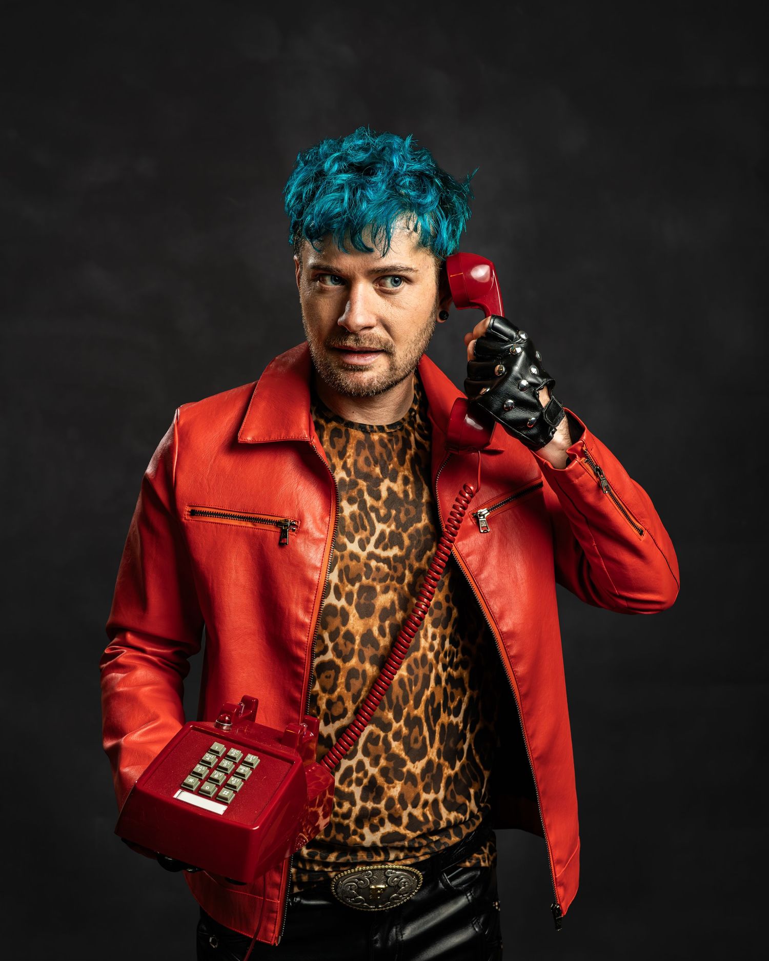Teal-haired performer in red leather jacket and leopard shirt holds a vintage red telephone prop.