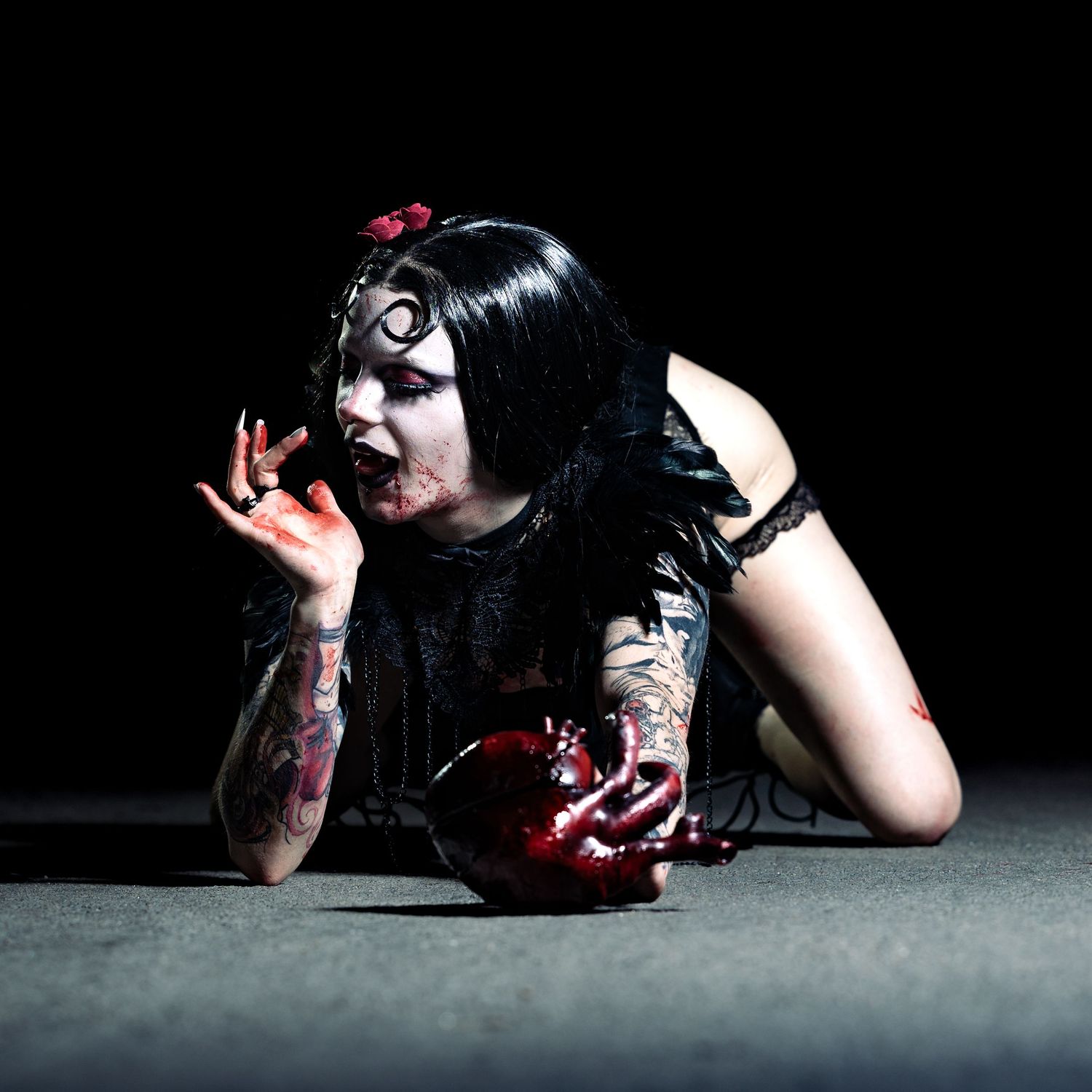 Gothic performer in dark makeup and costume poses dramatically on floor with theatrical blood effects.