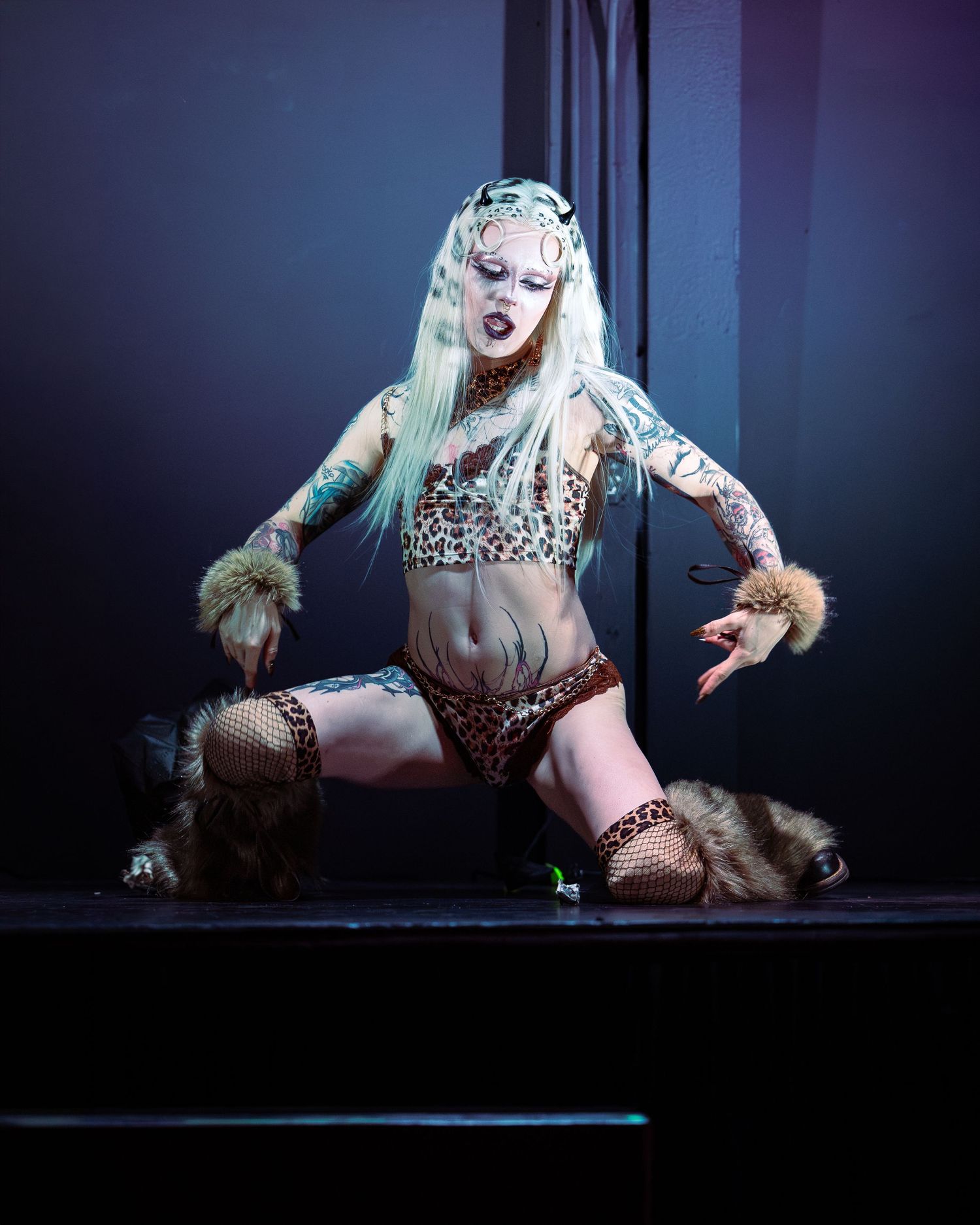 Tattooed performer in leopard print costume with fur accessories poses dramatically on stage under blue lighting.