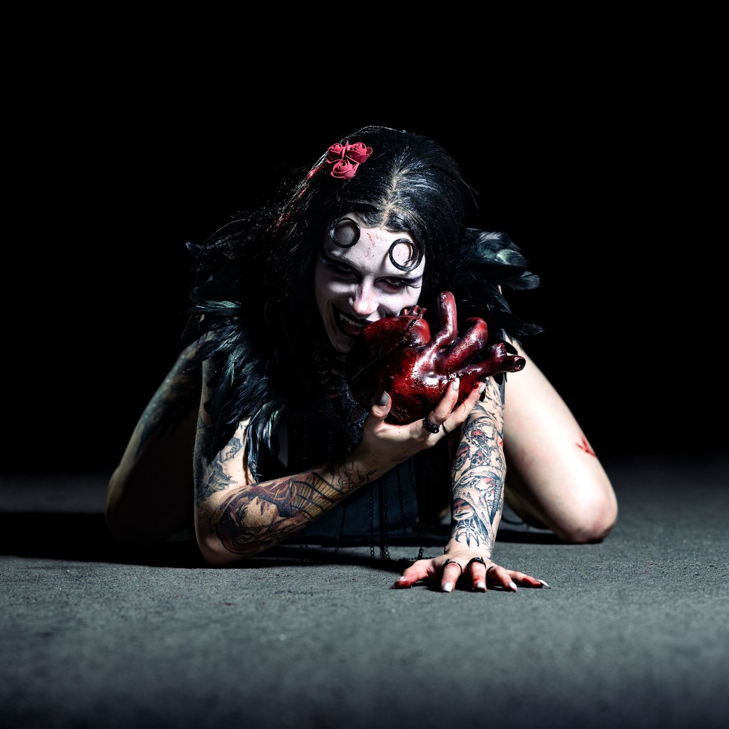 Tattooed woman in dramatic zombie Halloween makeup crawling on ground with bloody hand prop in dark setting.
