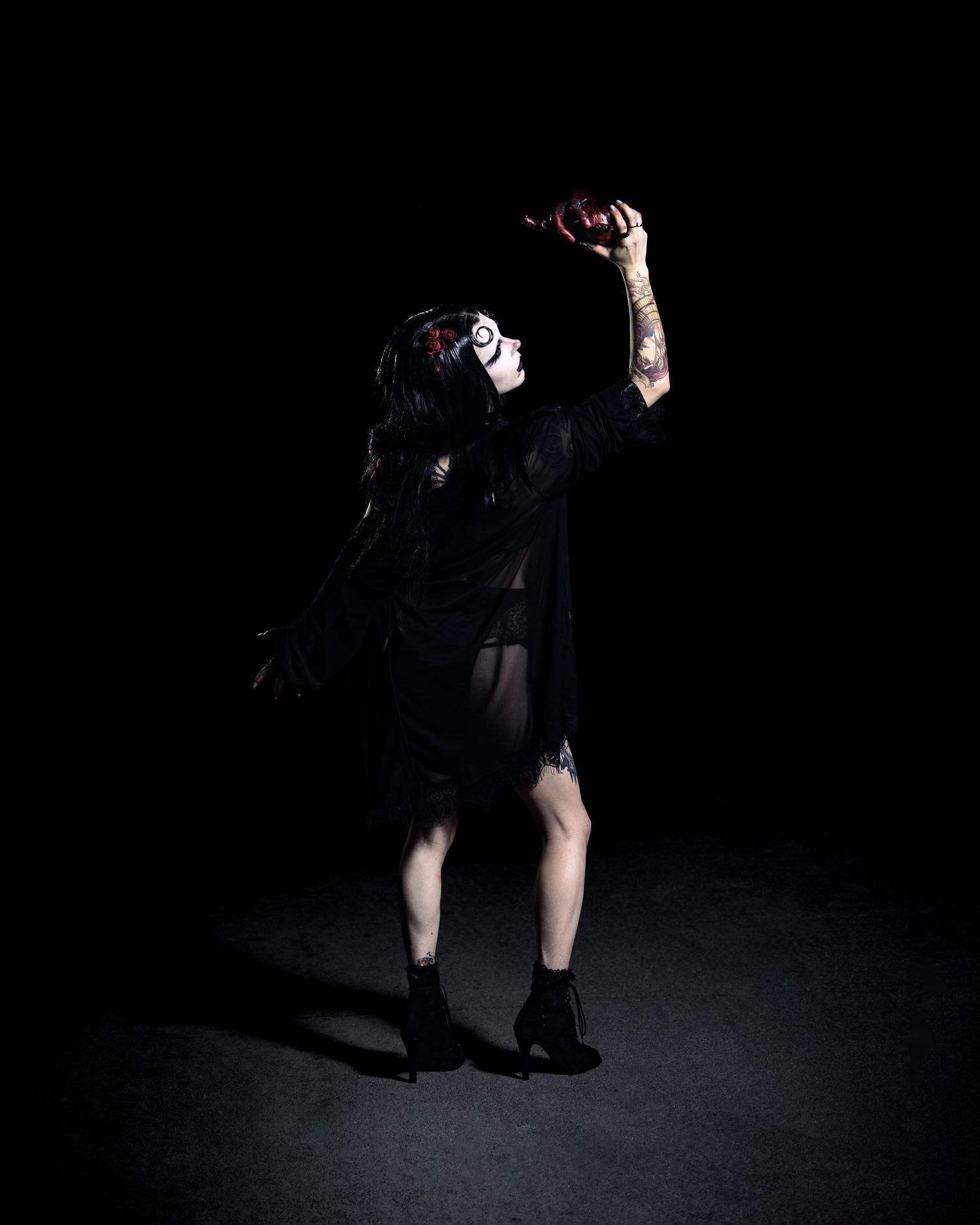Person in gothic makeup and black outfit holding a bottle aloft in dramatic dark studio lighting.