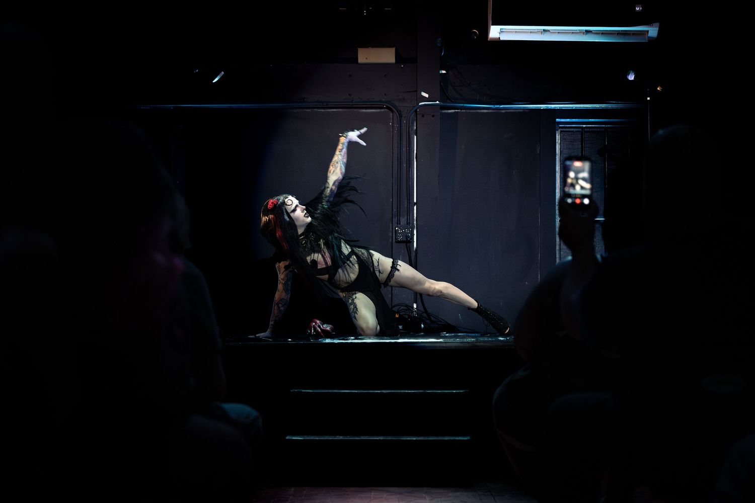 A dramatic performer in dark costume strikes a pose under stage lighting in a dimly lit venue.
