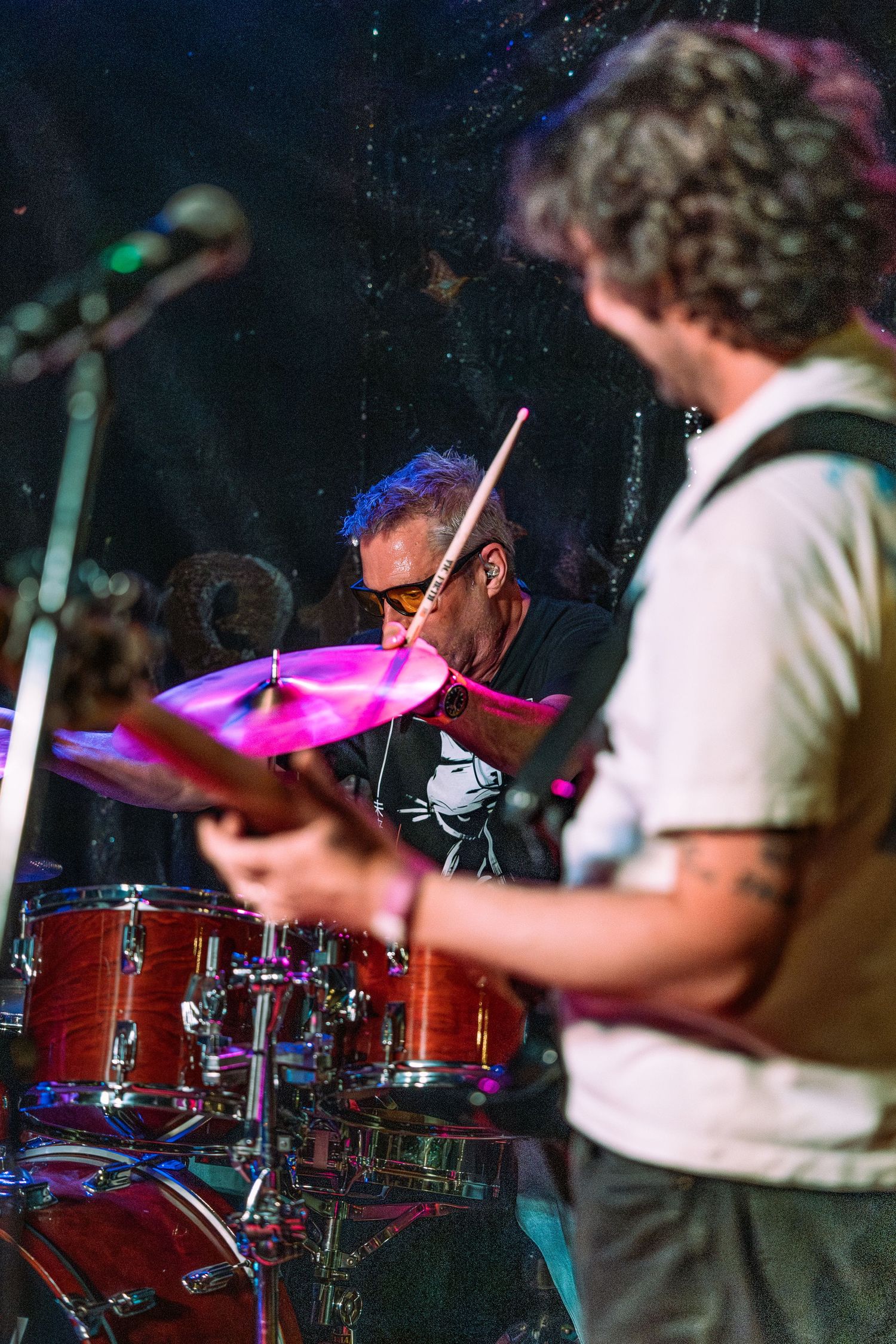 Drummer playing cymbals on stage with guitarist in foreground during live rock band performance.