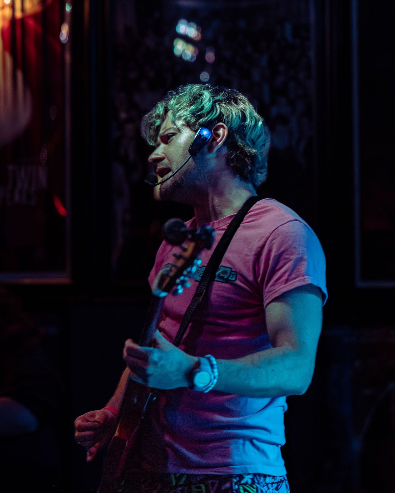 Male musician playing guitar on stage with headset microphone under blue and green stage lighting.