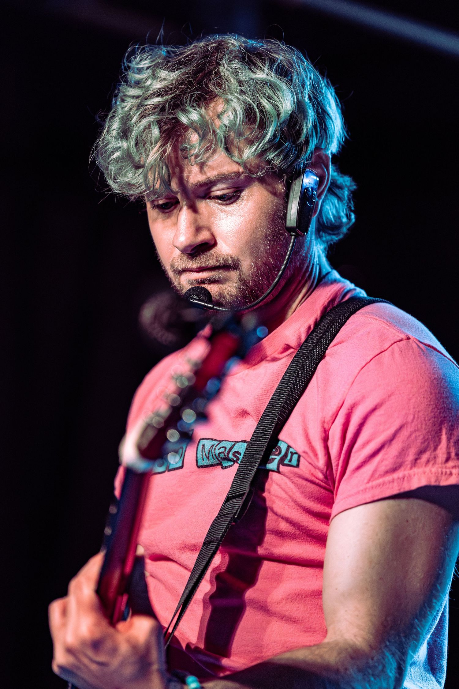 Curly-haired male guitarist performing on stage wearing a pink t-shirt and in-ear monitor.
