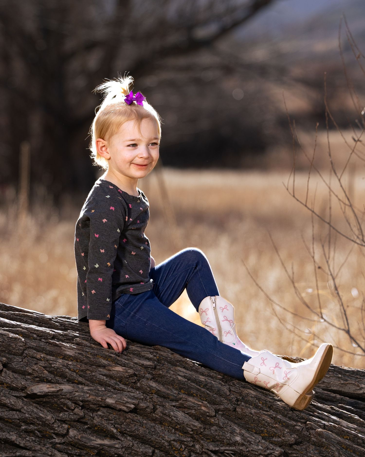 Smiling blonde toddler girl sitting on a log outdoors, wearing jeans, floral top, and pink cowboy boots.
