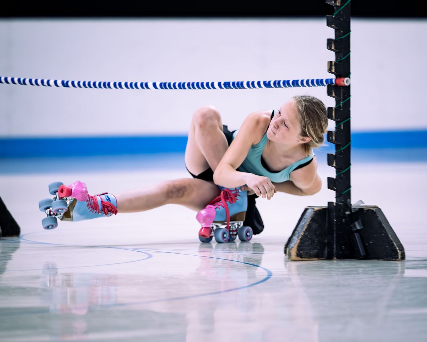 Woman in roller skates crouching near a rink barrier, wearing a teal top during an indoor skating session.