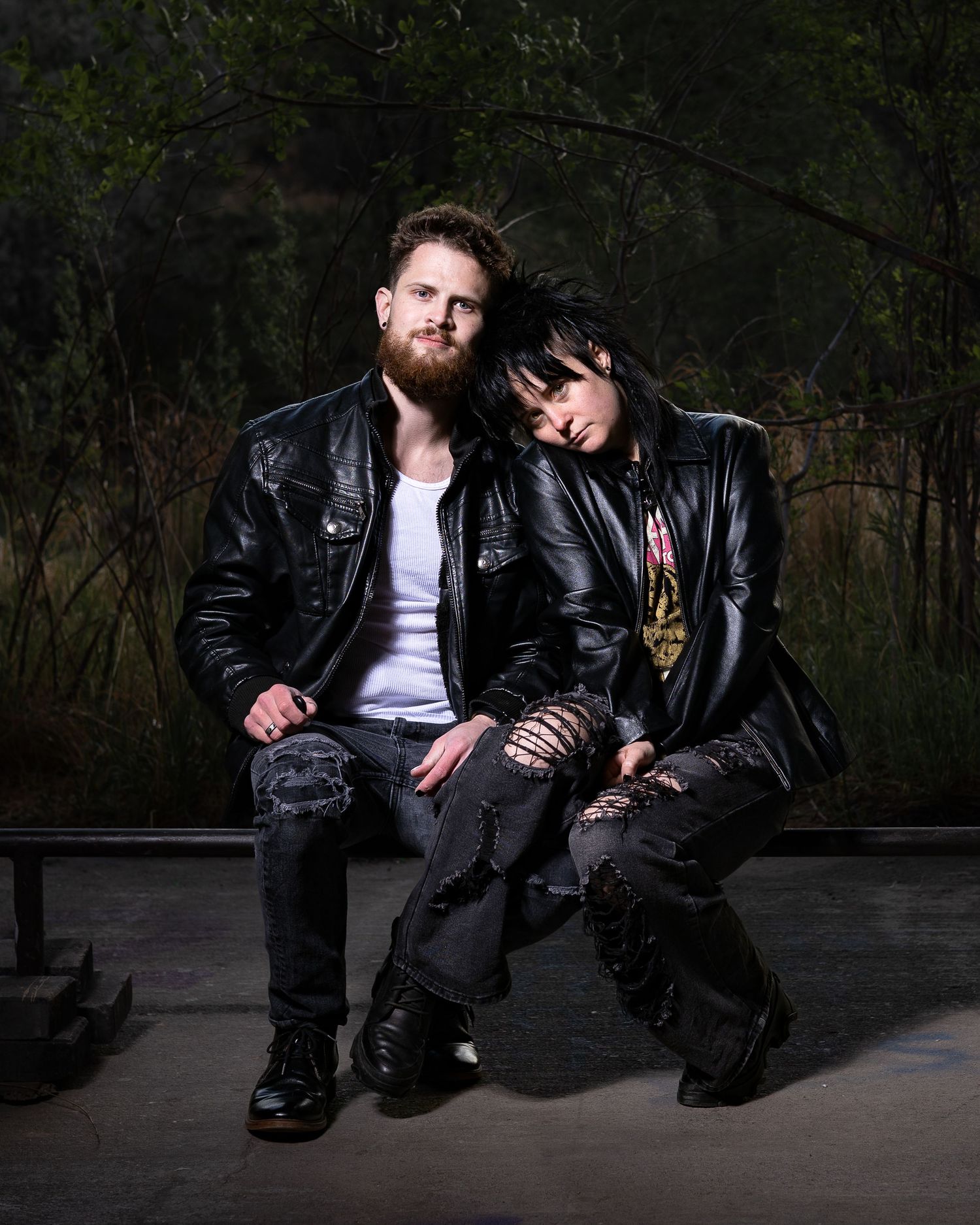 Two rock-style individuals in black leather jackets pose together outdoors in a moody, dramatic portrait setting.