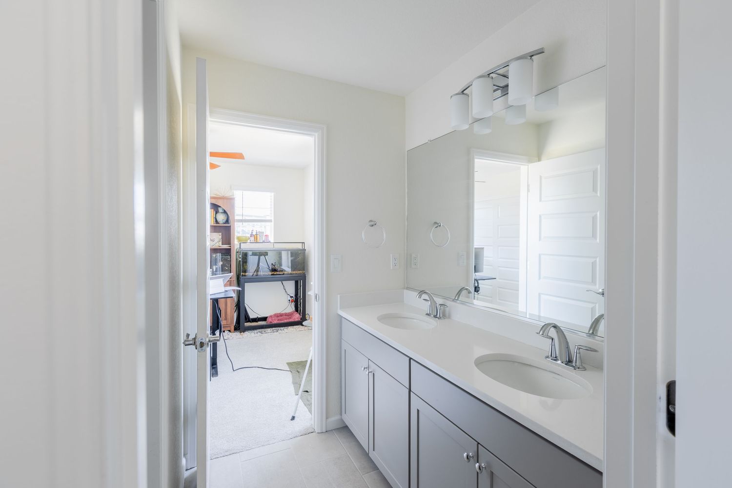 Bright bathroom with dual sink gray vanity, large mirror, and view into carpeted bedroom.