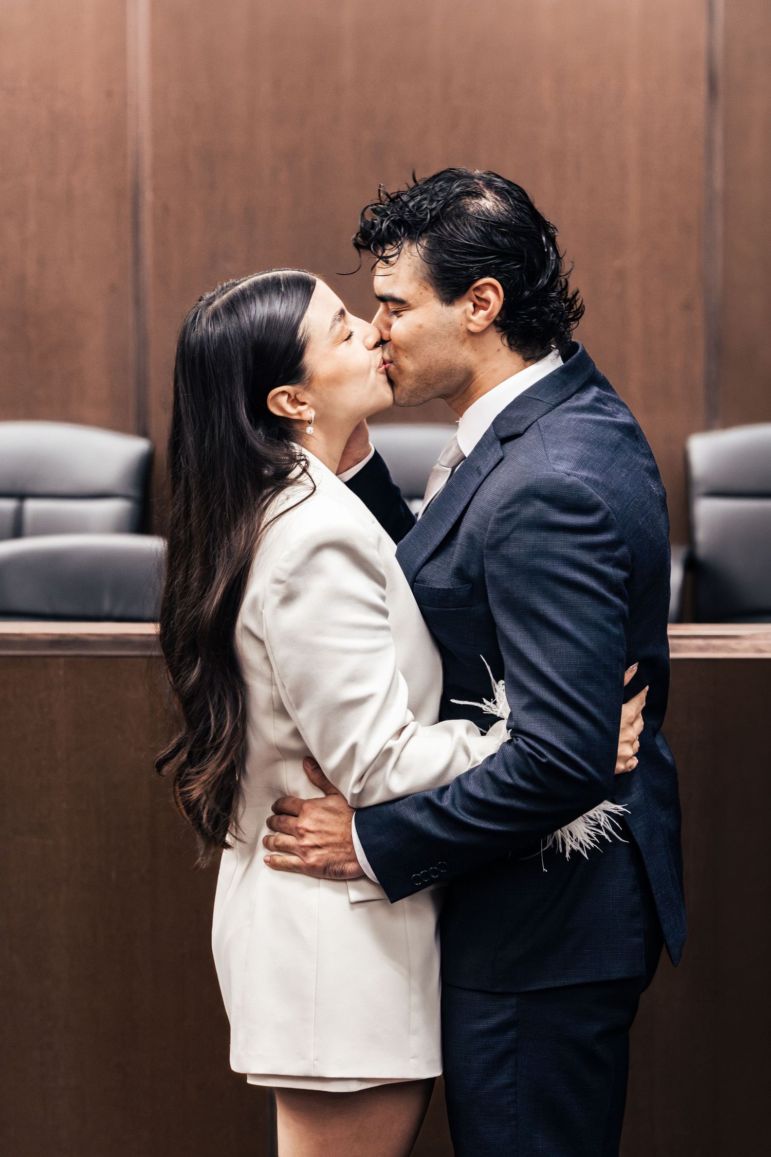 Couple sharing a romantic kiss in a courtroom, woman in white suit and man in navy blue suit embracing.