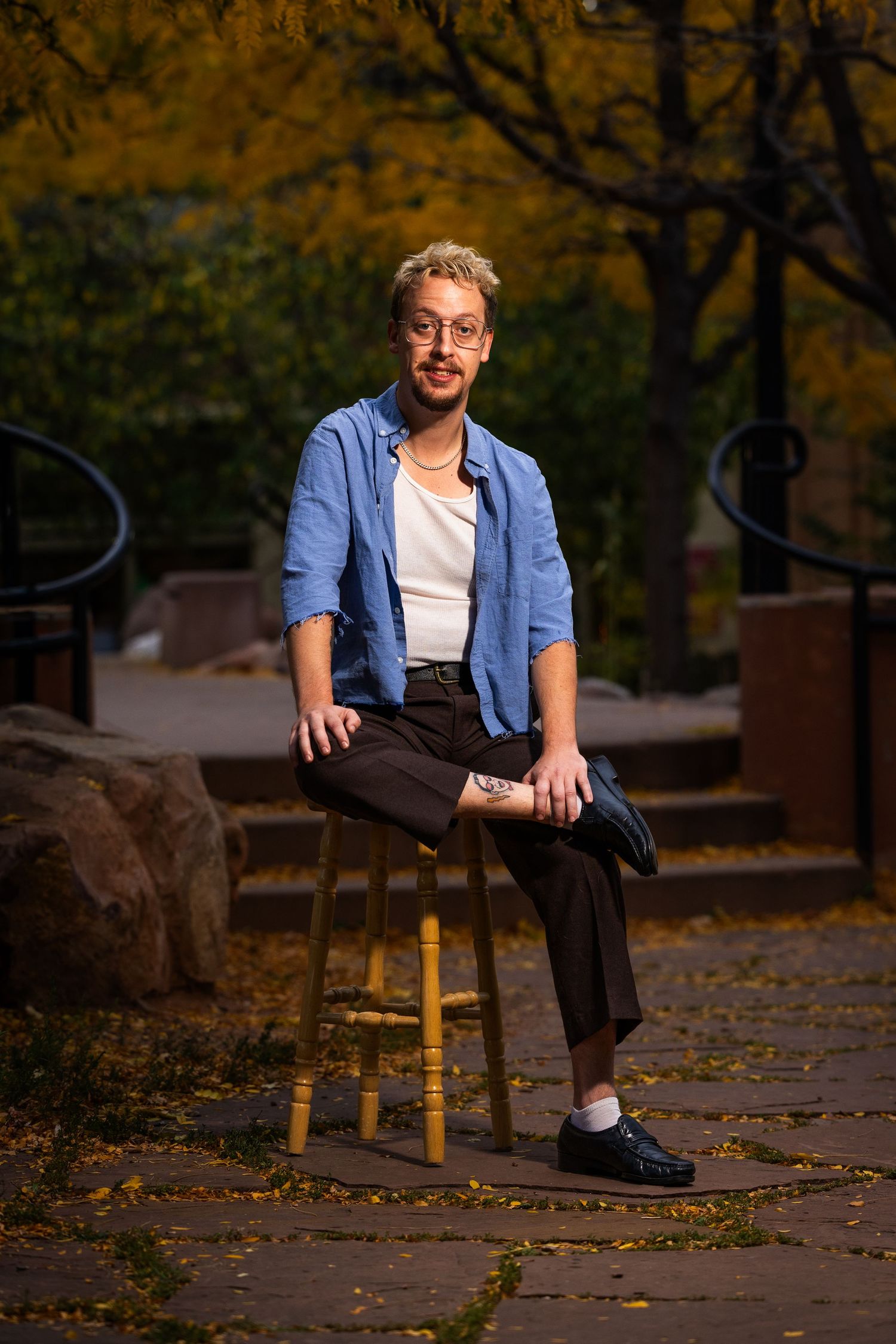 A person in a blue denim shirt and dark pants sits casually on a yellow post in an autumn outdoor setting.