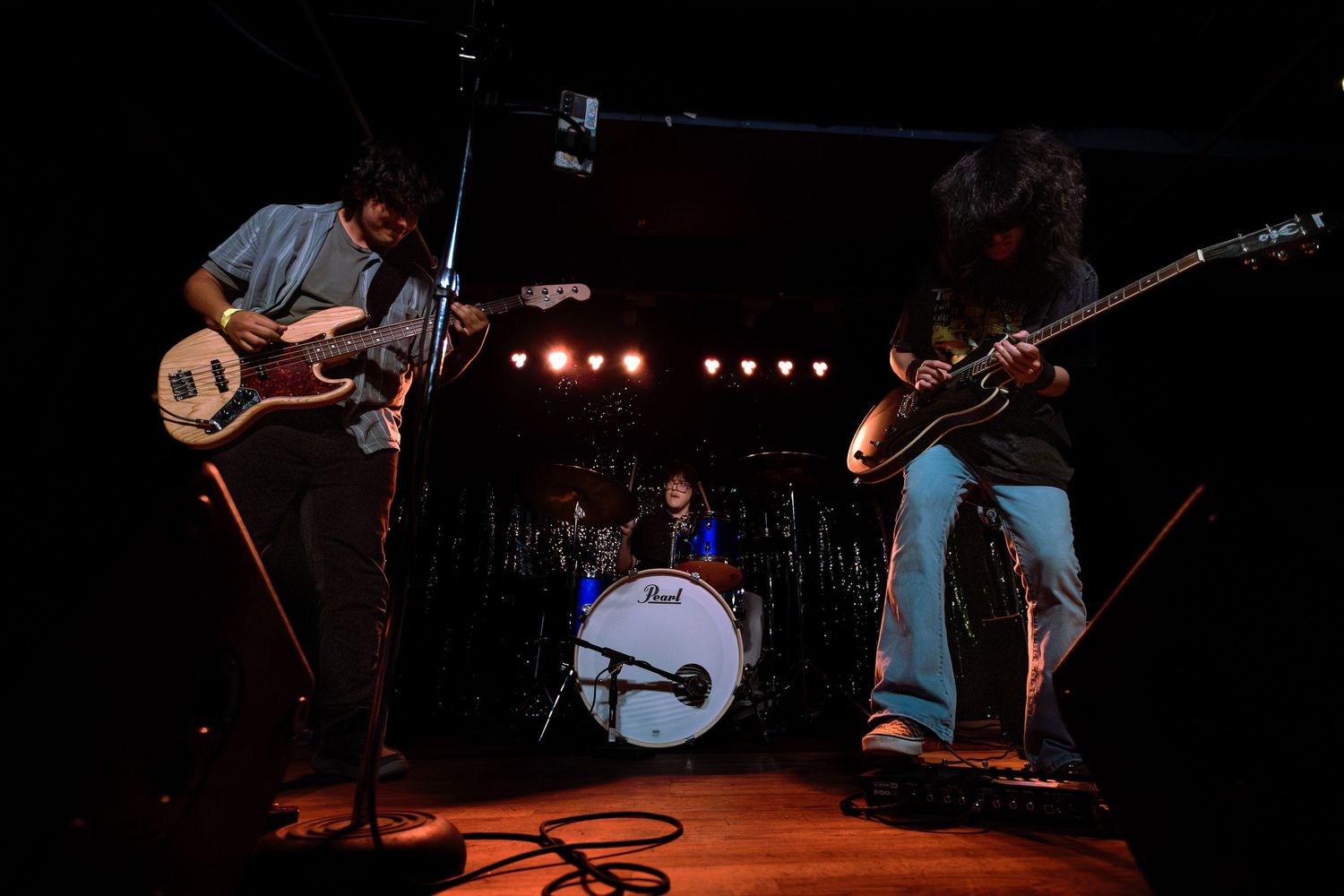 Three-piece rock band performing live on stage with guitars and Pearl drum kit under dramatic concert lighting.