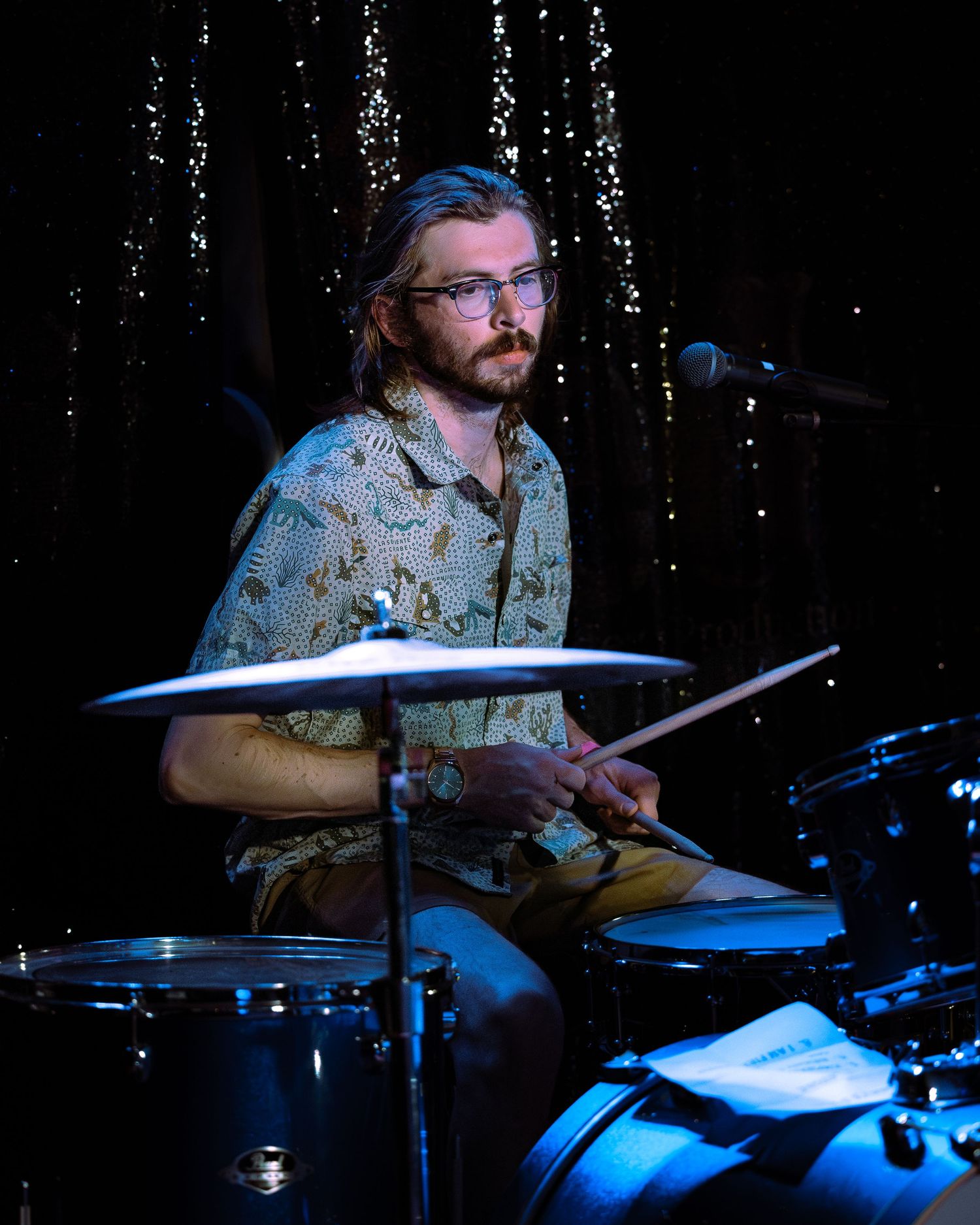 Bearded drummer with glasses holding drumsticks behind a drum kit on a dark stage.