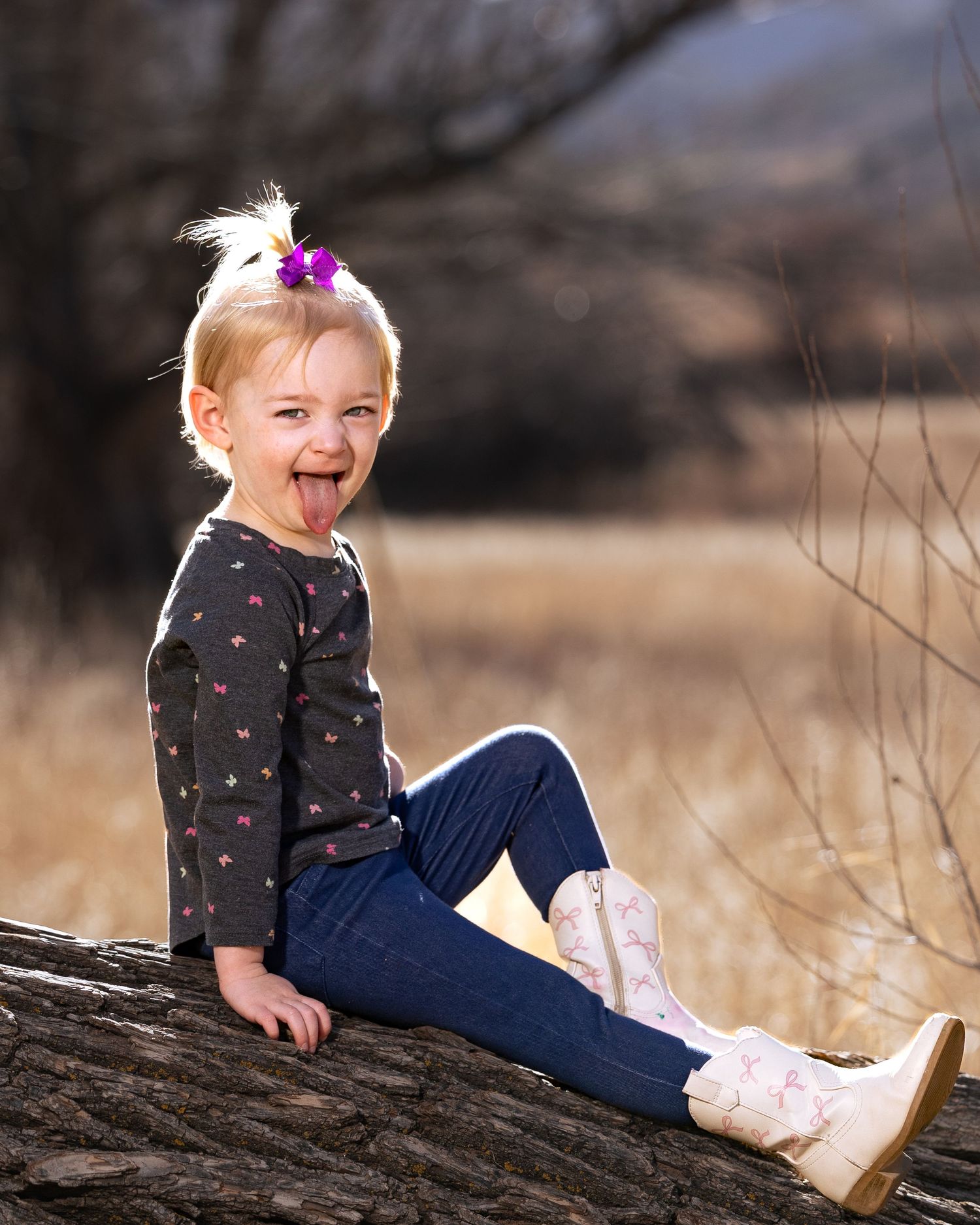 Cheerful blonde toddler girl sitting on a log, sticking out her tongue, wearing jeans and cowboy boots outdoors.