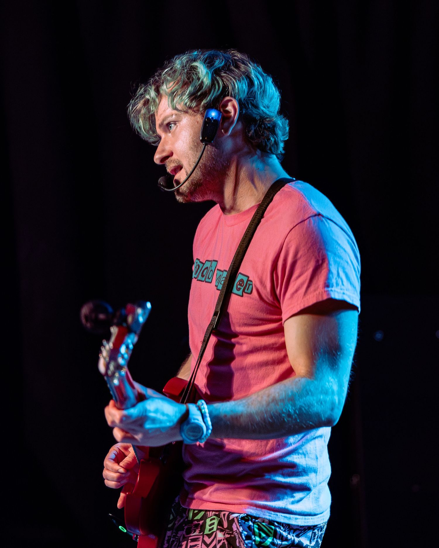 Young man playing red guitar on stage wearing pink t-shirt and headset microphone.