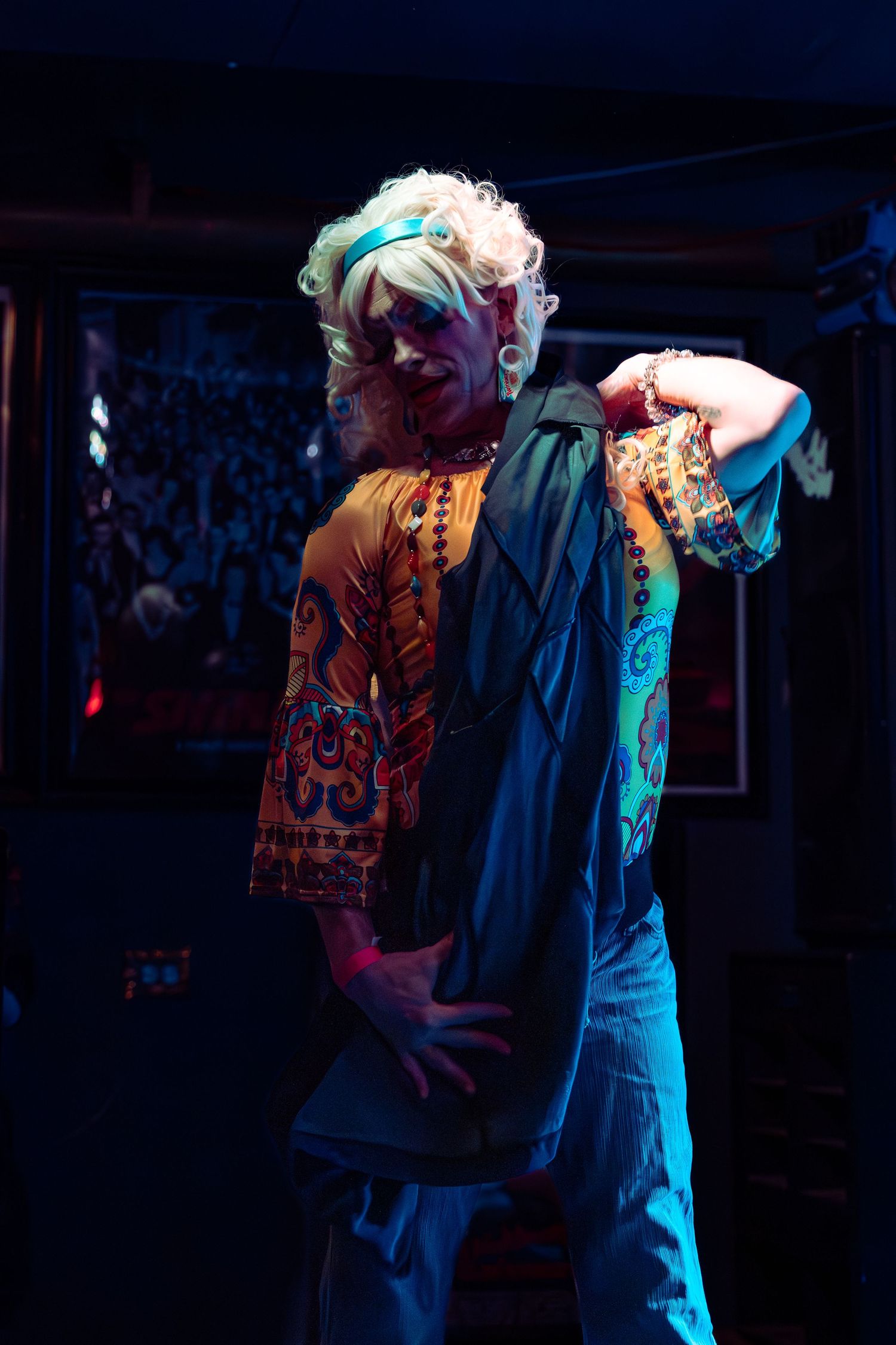 Performer with blonde curly hair in colorful bohemian shirt posing on dark stage under blue lighting.